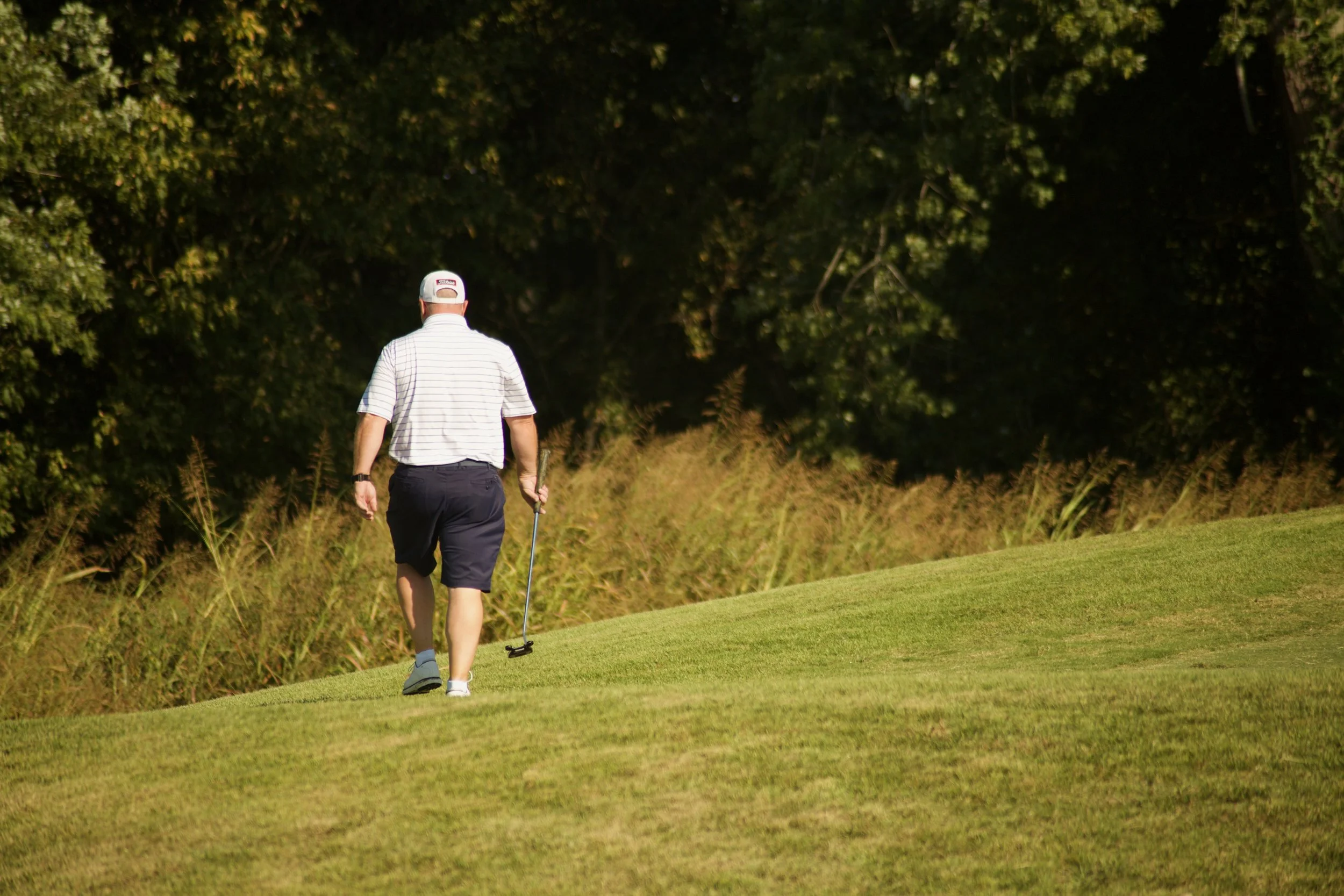 A man wearing a white striped polo, black shorts, and a white cap walking on a golf course, holding a golf club, with Trees and grass in the background.