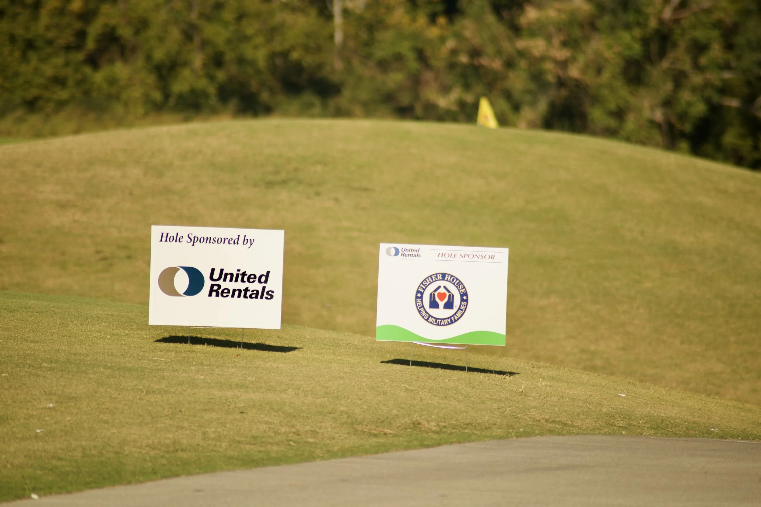 Golf course with yellow flag in the background and sponsor signs on the grass in the foreground, including United Rentals and Fisher House logos.