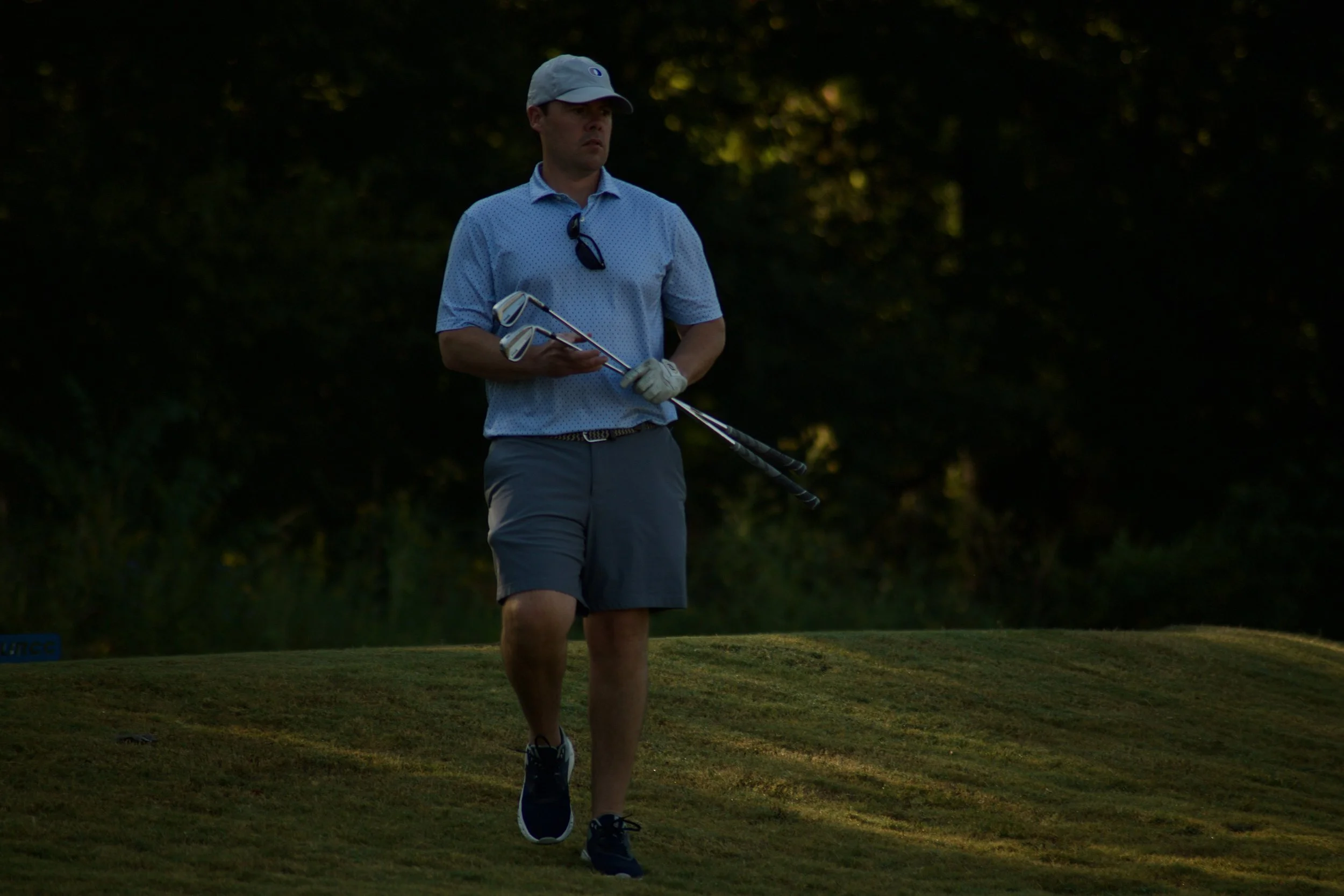 A man in golf attire standing on a golf course holding three golf clubs with sunglasses hanging from his shirt collar, surrounded by trees.