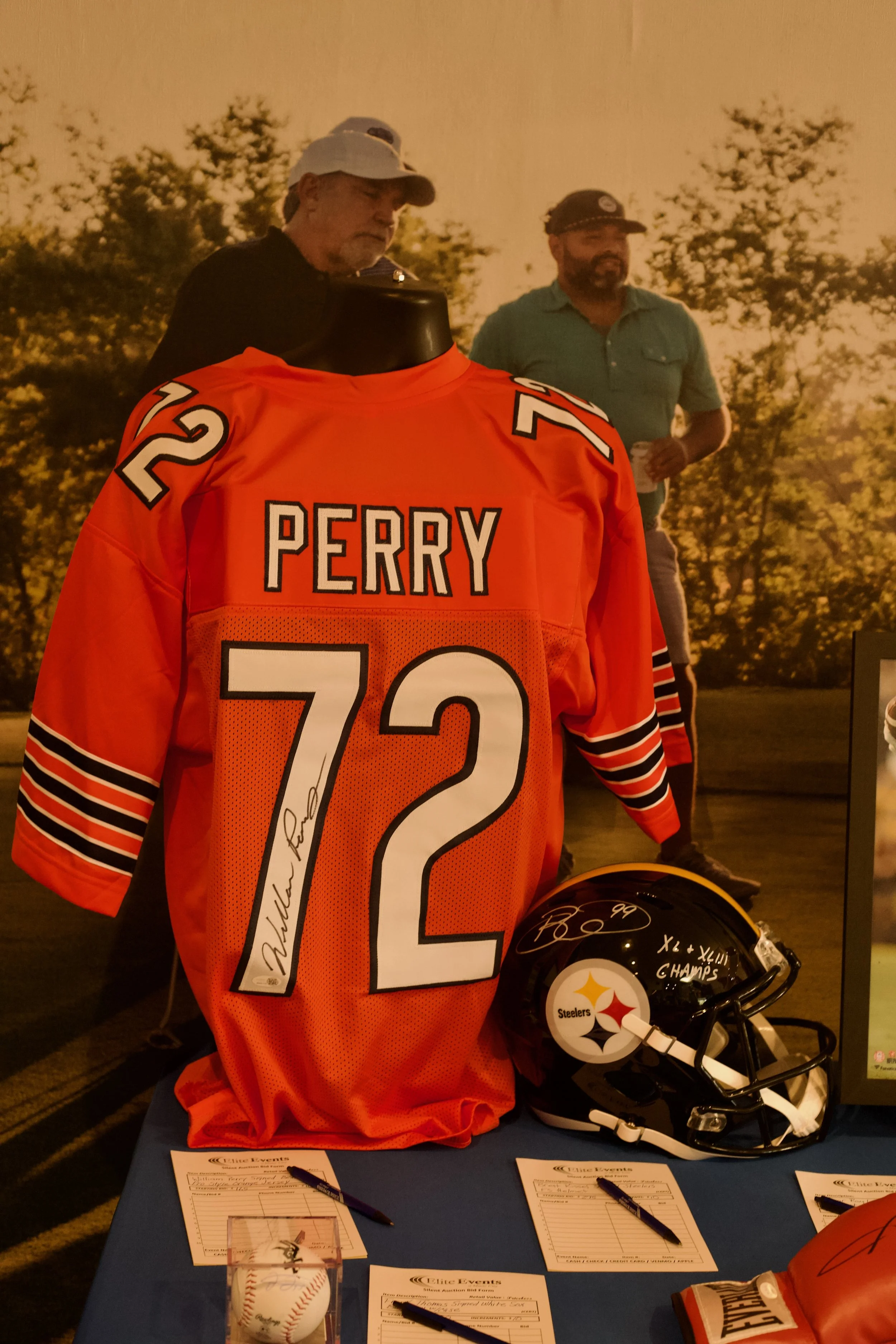 A display of an orange football jersey with the name 'PERRY' and the number 12, signed by a player. Nearby are signatures on a football helmet and football, along with papers for signing, in a memorabilia setting.