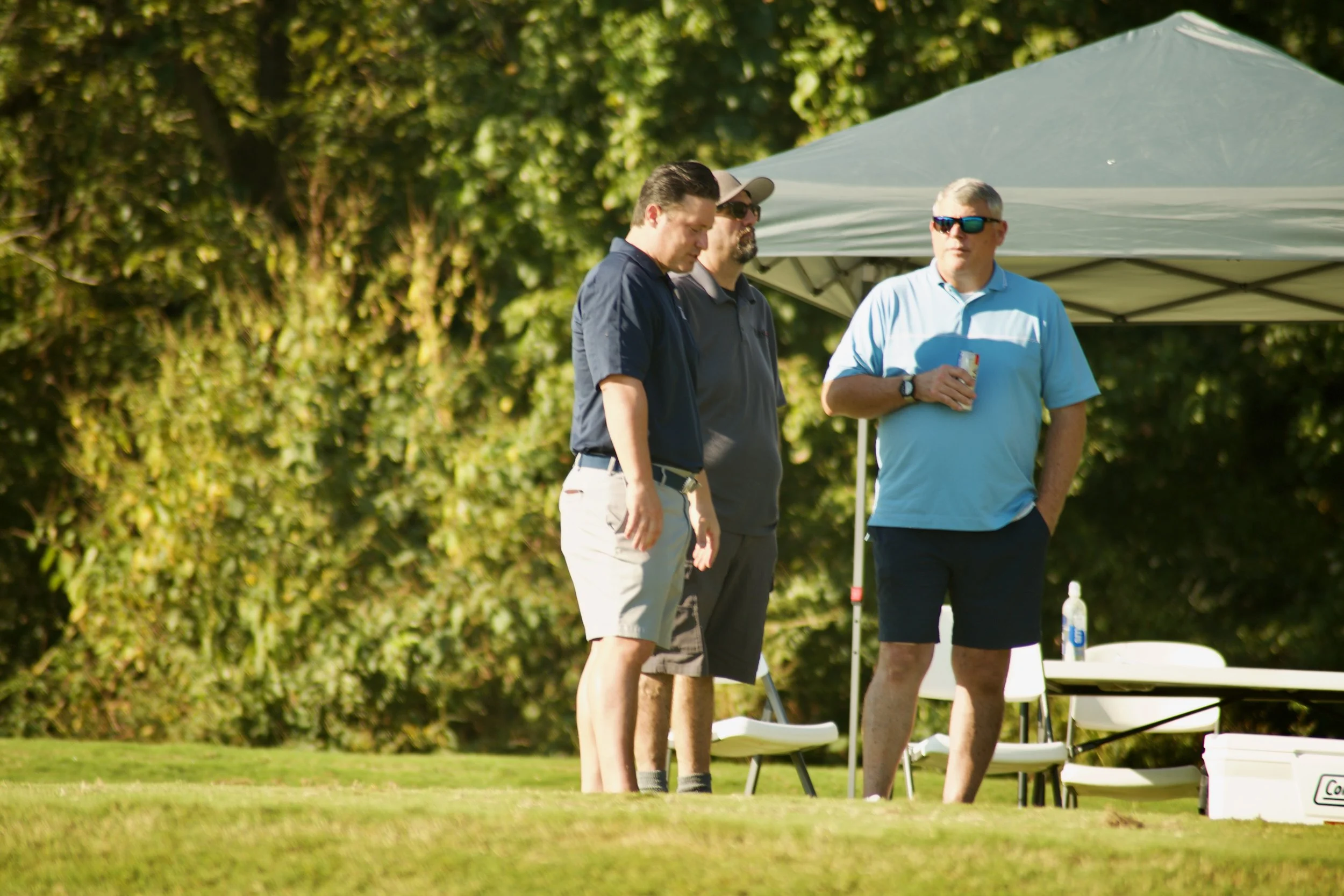 Four men standing outdoors near a table under a canopy, with a background of green trees, in sunlight.