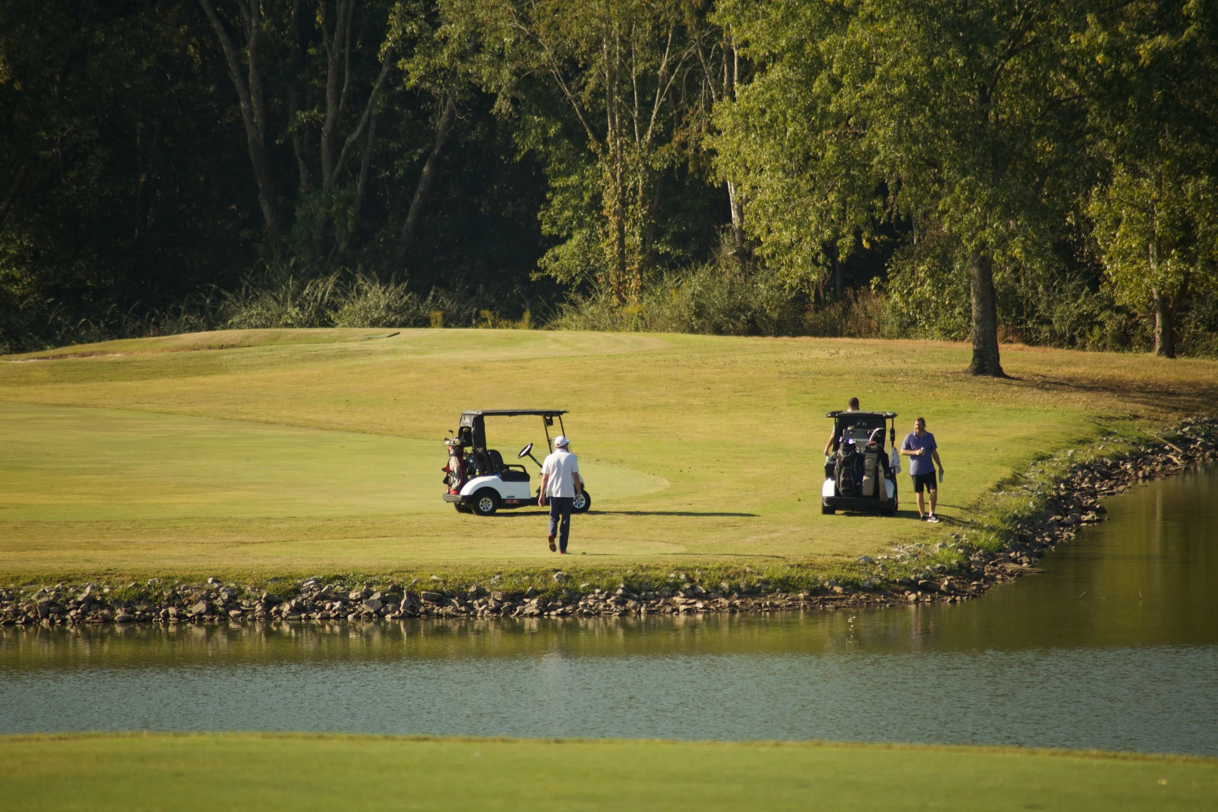 Golf course with three golfers near water, surrounded by trees.