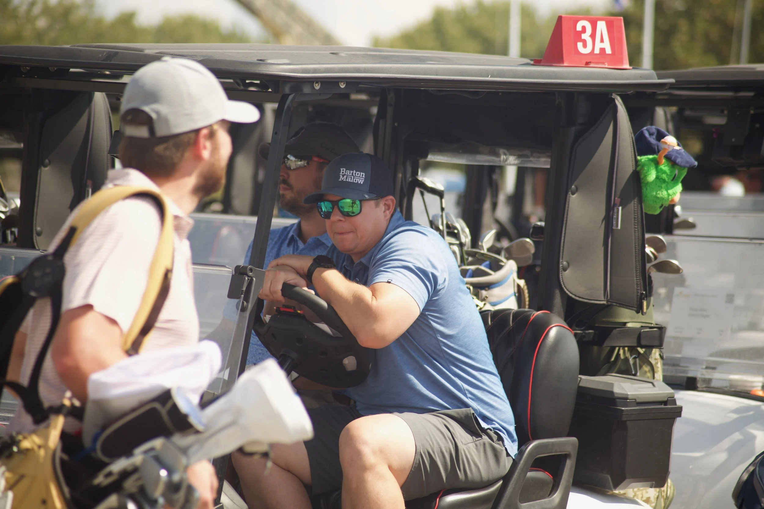 A man sitting in a golf cart with a blue shirt, sunglasses, and a cap, talking to a person standing nearby, with a golf bag and clubs attached to the back.
