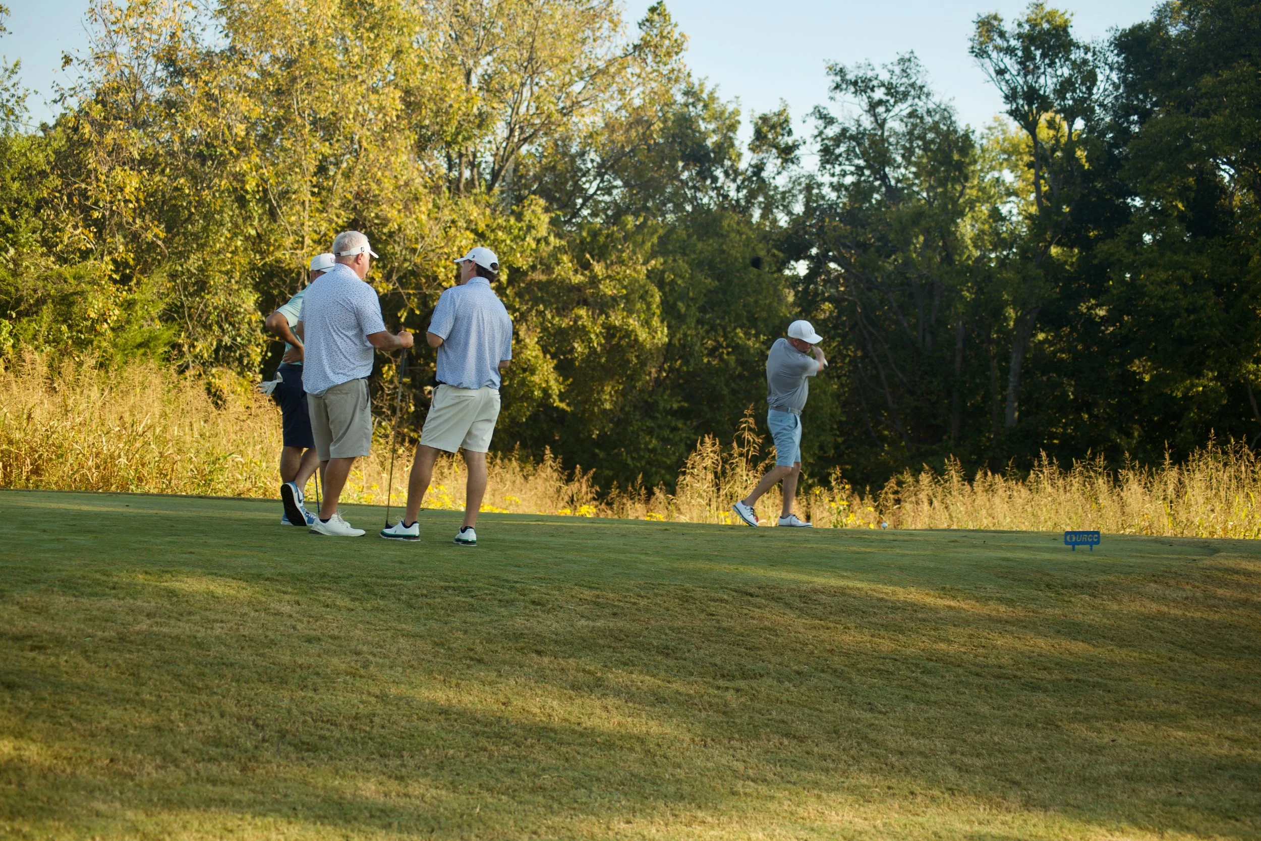 A group of six men playing golf on a lush green course surrounded by trees in the background, with one man preparing to take a shot.