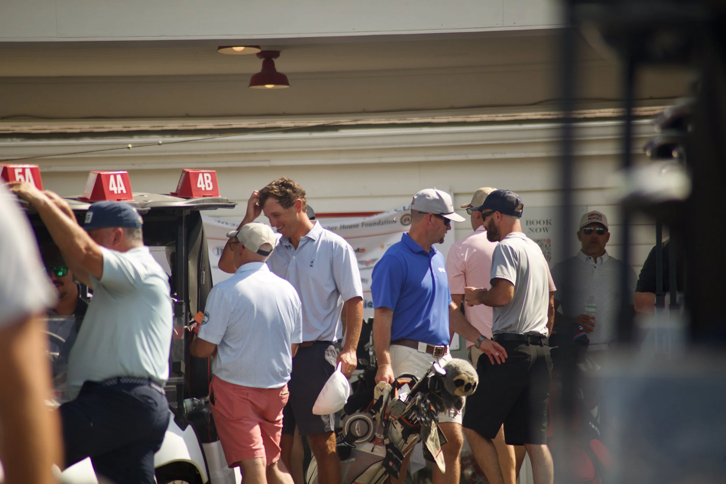 Group of people at a golf event, some wearing golf attire and hats, surrounded by golf bags and equipment, located outdoors near a building with a covered area.