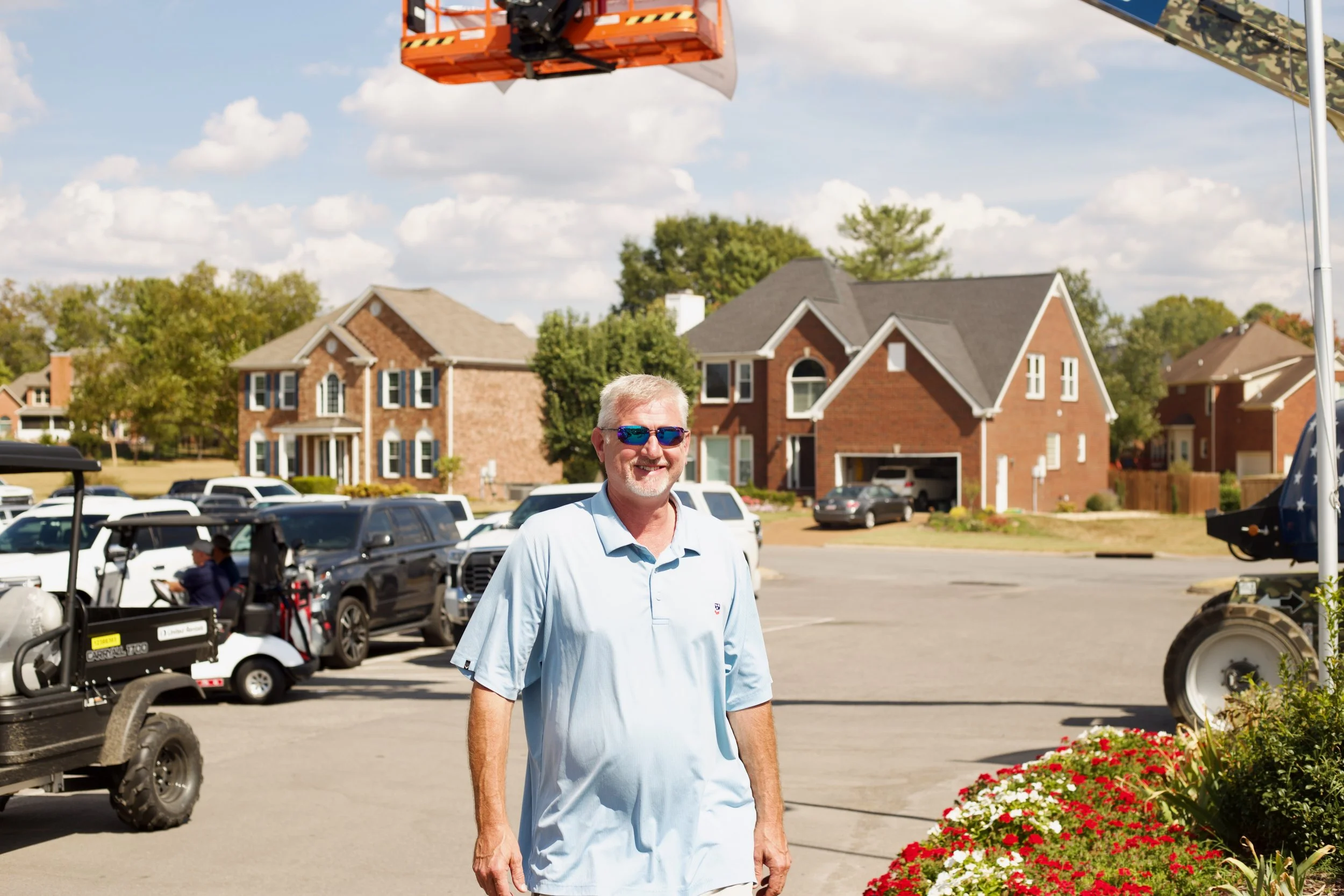 A man wearing sunglasses and a light blue polo shirt walking outdoors on a sunny day with houses, cars, and a golf cart in the background.