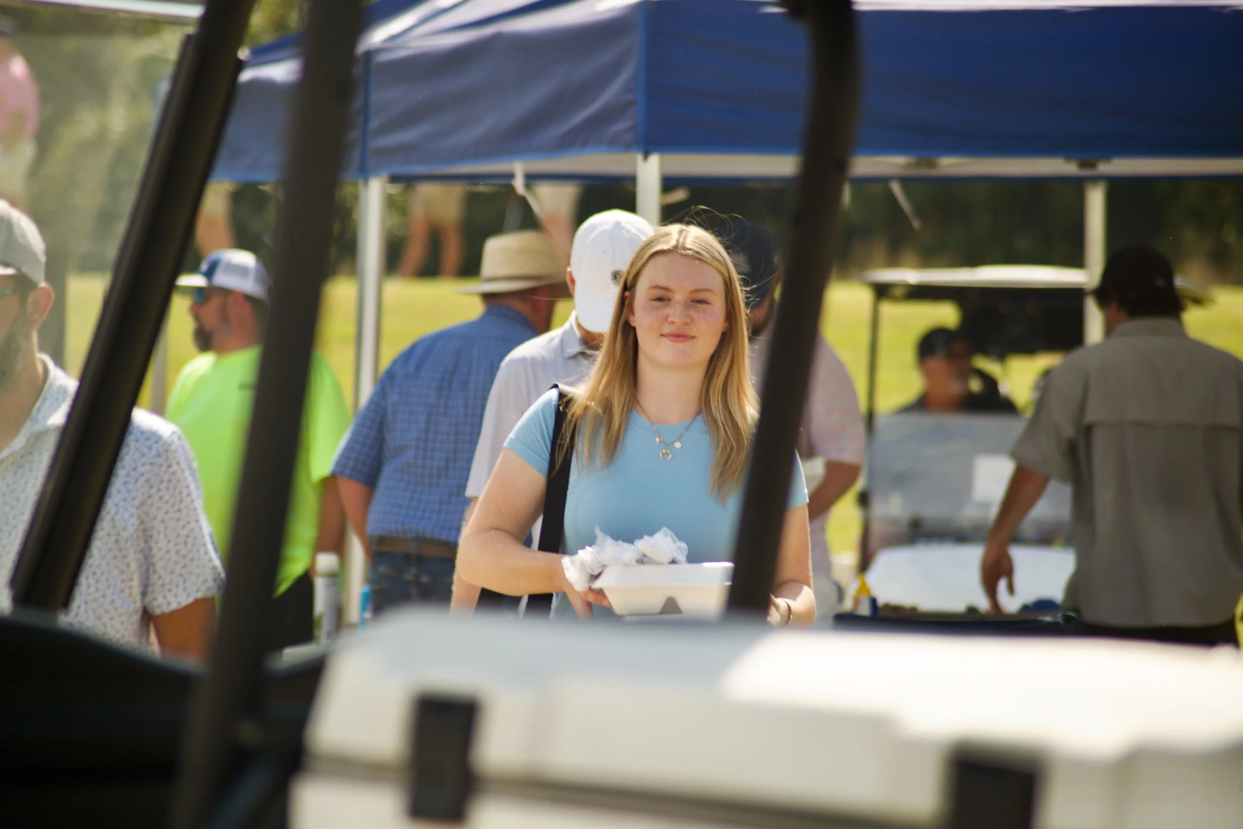 A young woman with blonde hair wearing a light blue shirt and a backpack standing in line at an outdoor event, holding a tray with food. Several people are visible in the background under a blue tent, and the photo appears to be taken from inside a v