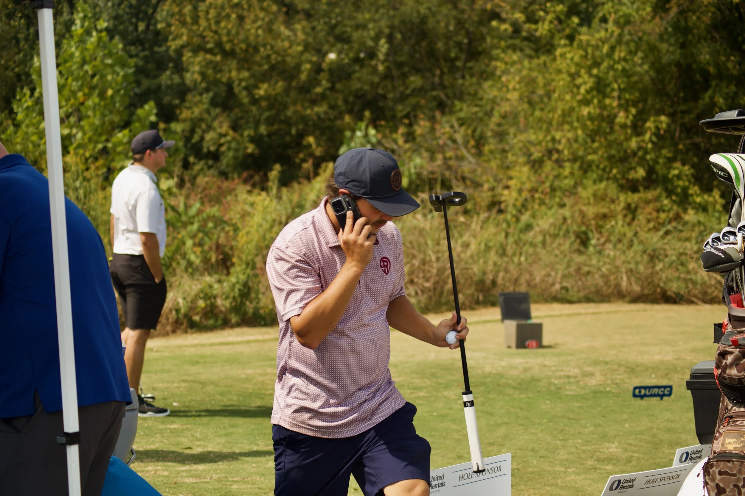 A man in a pink checkered shirt and dark shorts is holding a golf club and talking on his cellphone at an outdoor golf event. Two other men are visible in the background, one wearing a white shirt and black cap, the other wearing a blue jacket. There