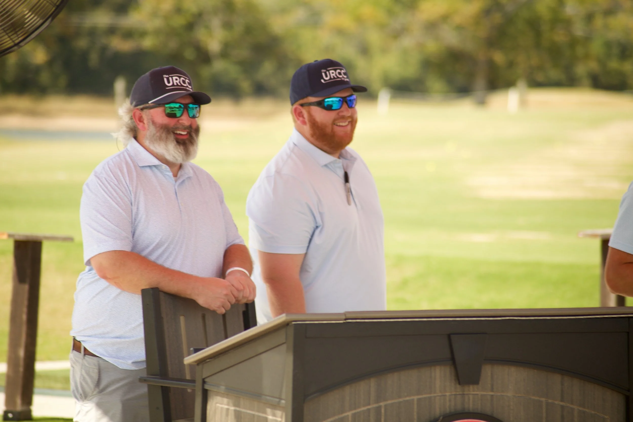Two men wearing sunglasses and hats with 'URCC' logo, standing outdoors near a keyboard or organ, smiling and enjoying a sunny day in a park with trees in the background.