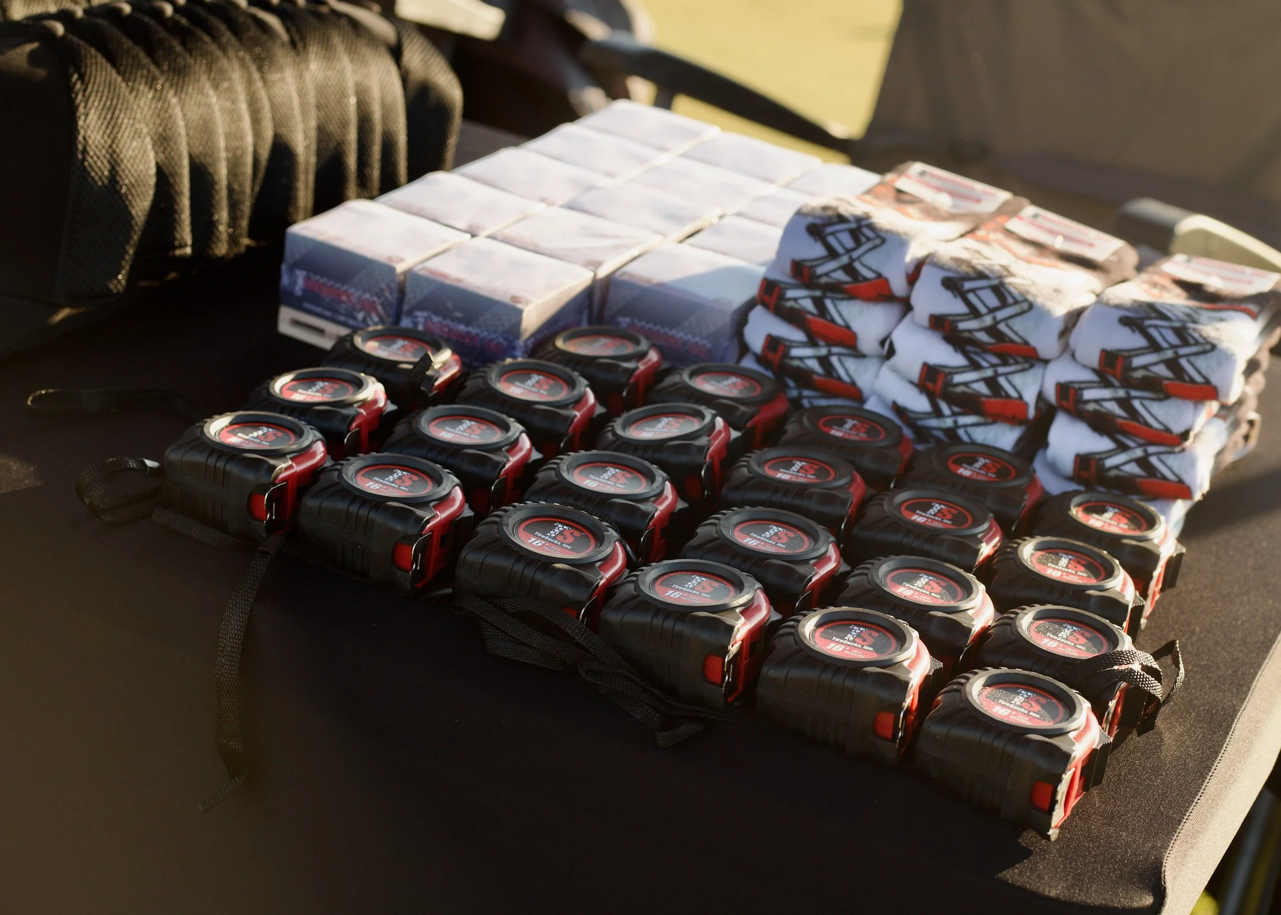 A table with multiple black and red measuring tapes, boxes, and folded shirts or cloth with a logo on it, set outdoors.