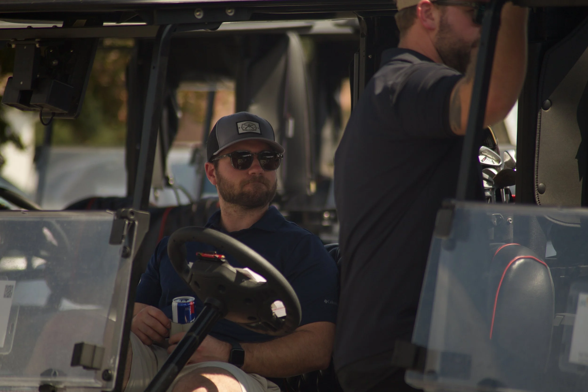 Two men sitting inside a golf cart. One man is wearing sunglasses and a hat, holding a soda can. The other man is standing, adjusting the roof of the golf cart.