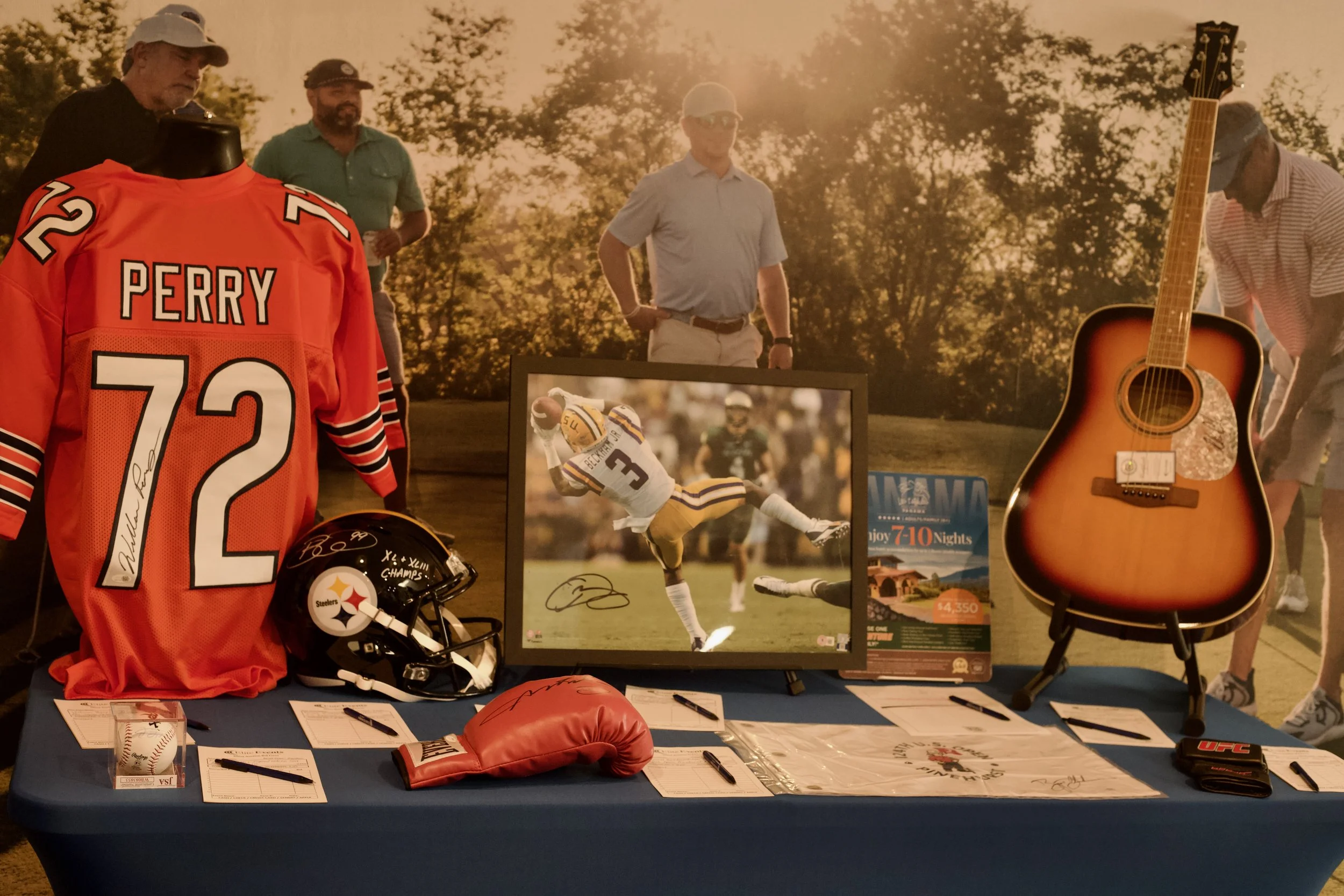 Display of sports memorabilia with an orange football jersey signed by Terry Perry, a black football helmet with the Pittsburgh Steelers logo, a boxing glove, a framed autographed photo of a football player, a guitar, and various signatures and poste
