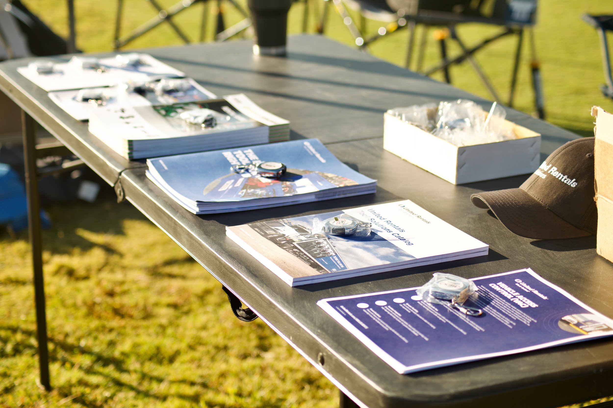 A black outdoor table with multiple brochures, magazines, and promotional materials about rental cars, along with a car key and a cap, set up on a grassy area during the daytime.