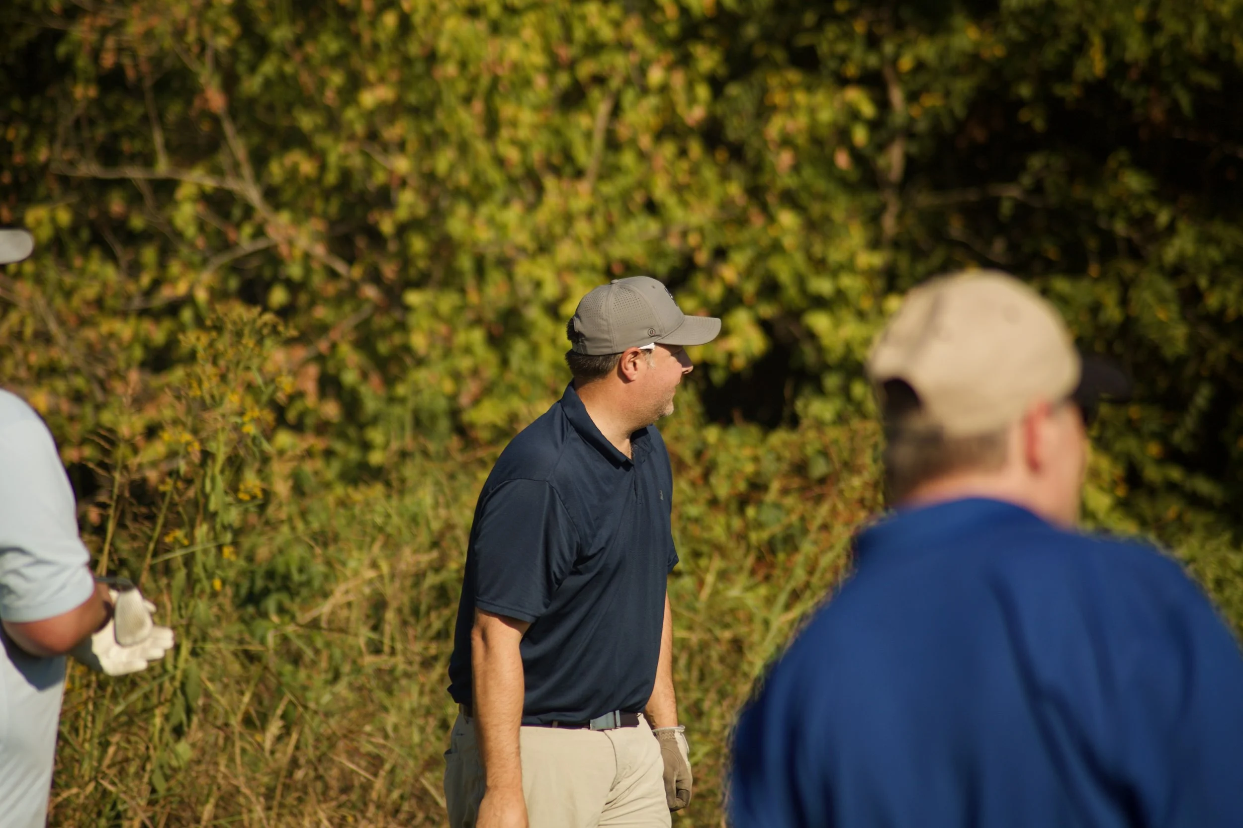 A man wearing a gray cap and dark shirt stands outdoors with a group of people, with a green leafy background.