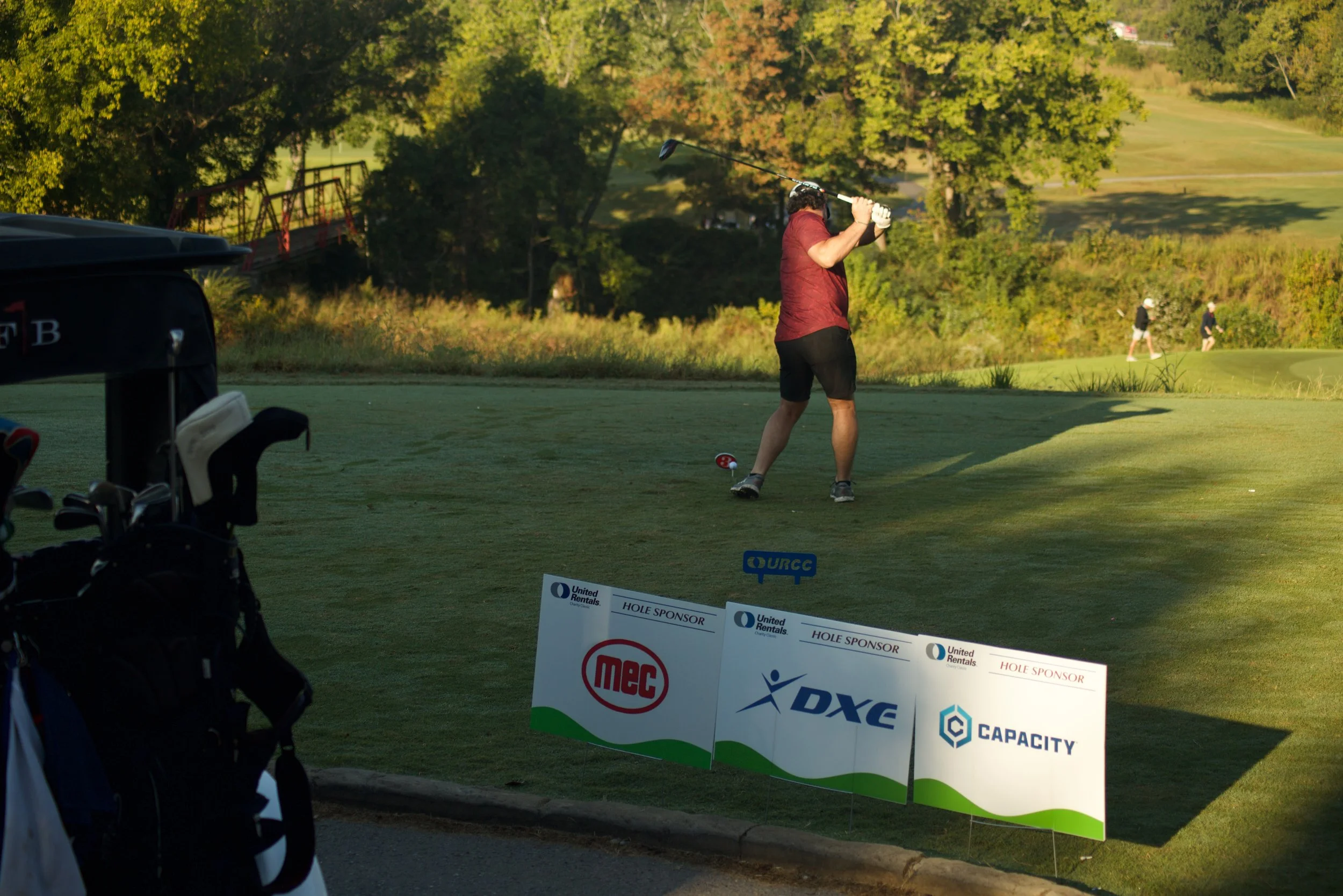 A man in a red shirt and black shorts playing golf swings a club on a golf course. There are trees in the background, and other people can be seen walking on the course. A golf bag with clubs and a sign with sponsor logos are in the foreground.