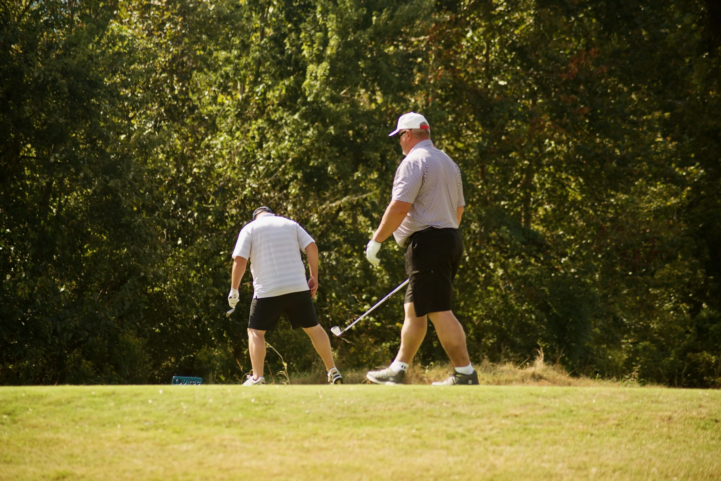 Two men on a golf course, one in a white polo and black shorts and the other in a white patterned shirt, black pants, and a white cap, preparing for a golf shot near a green surrounded by trees.