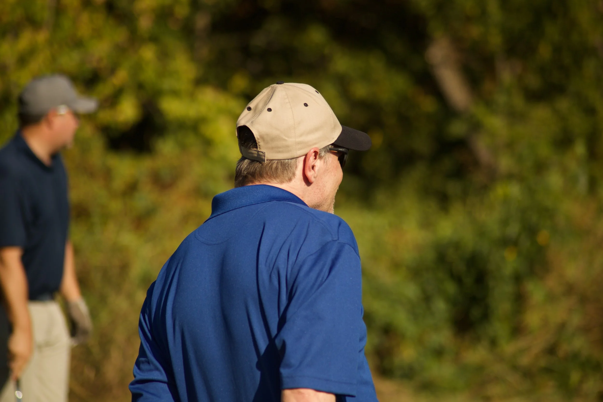 Two men outdoors, wearing caps and casual clothing, with a background of green trees and sunlight.