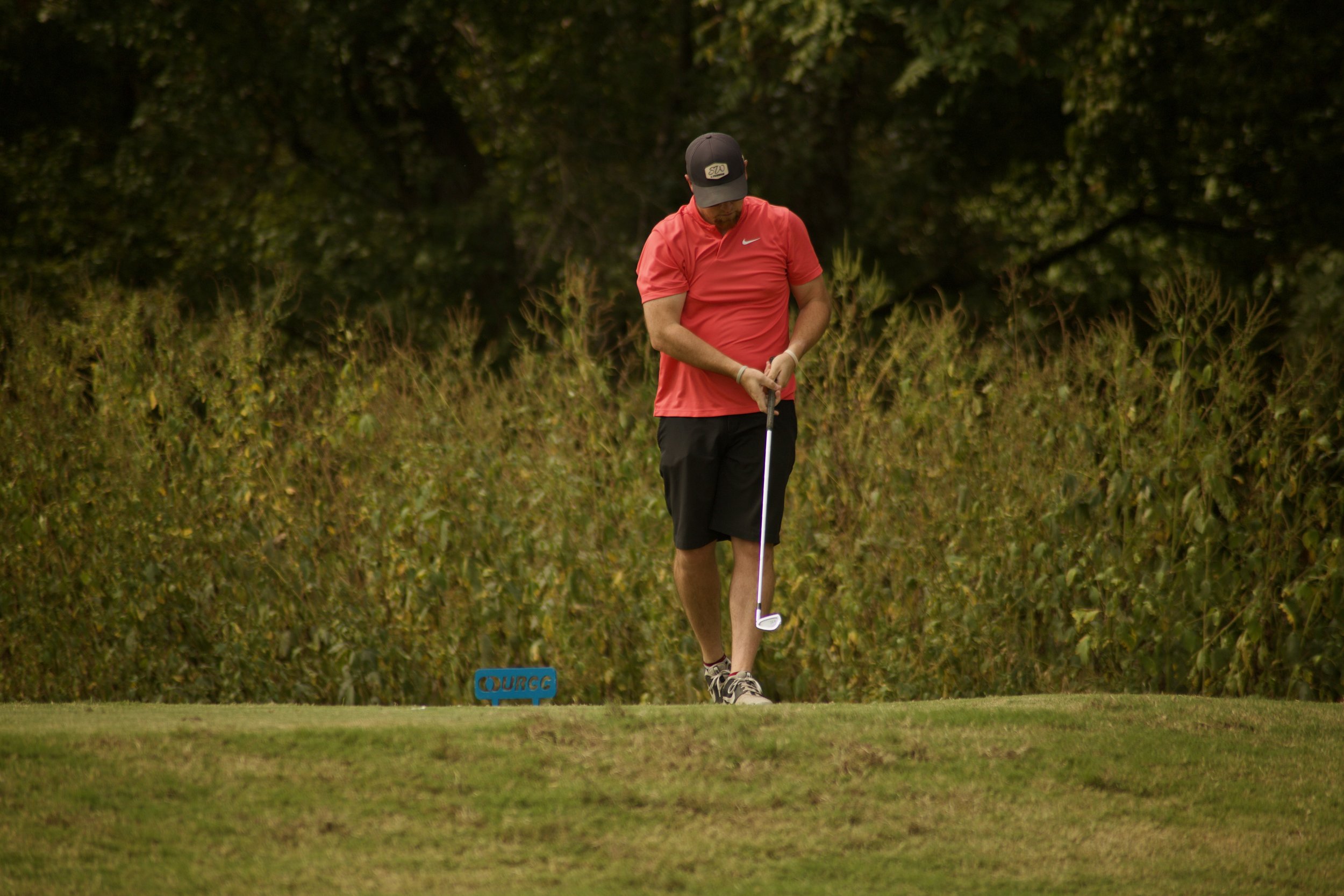 Man in red Nike shirt and black shorts playing golf on a grassy course, with a sign reading 'OUREC' nearby and trees in the background.