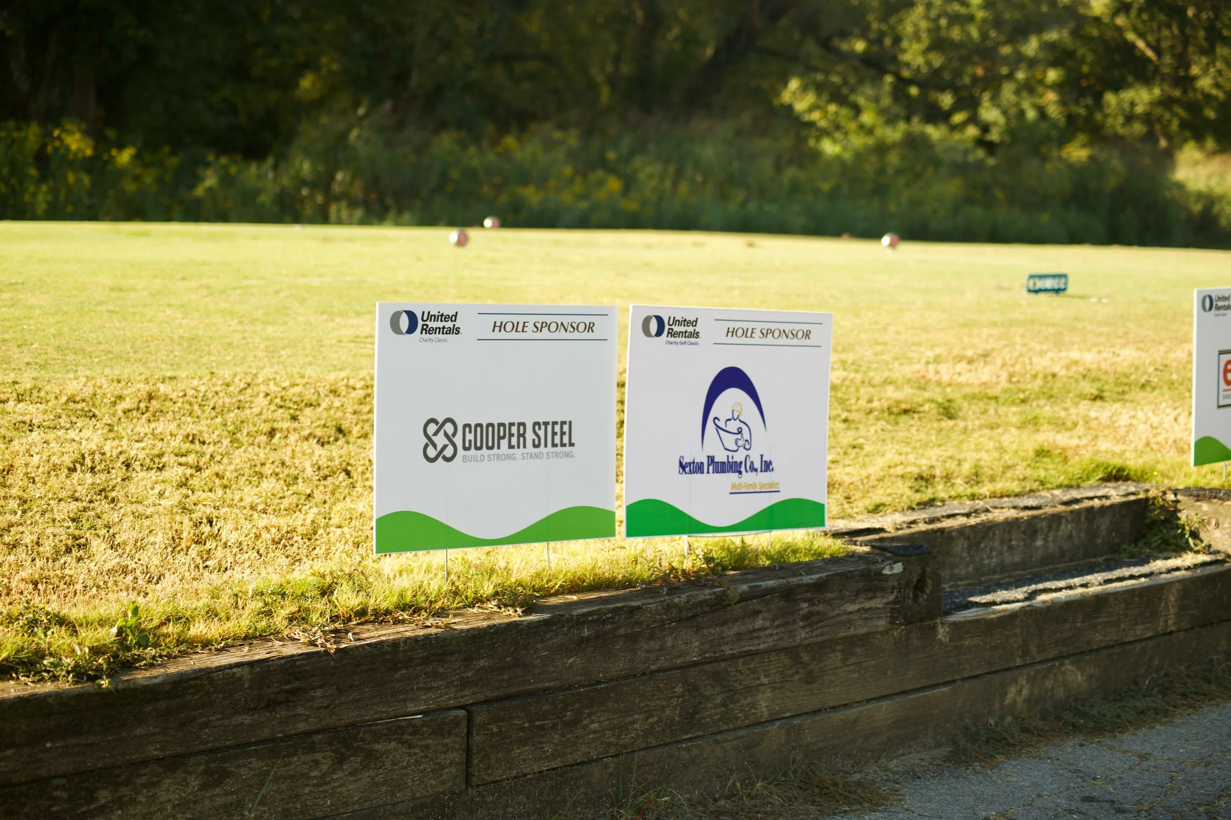 Signs with sponsors and local businesses on a golf course, with grass and trees in the background.