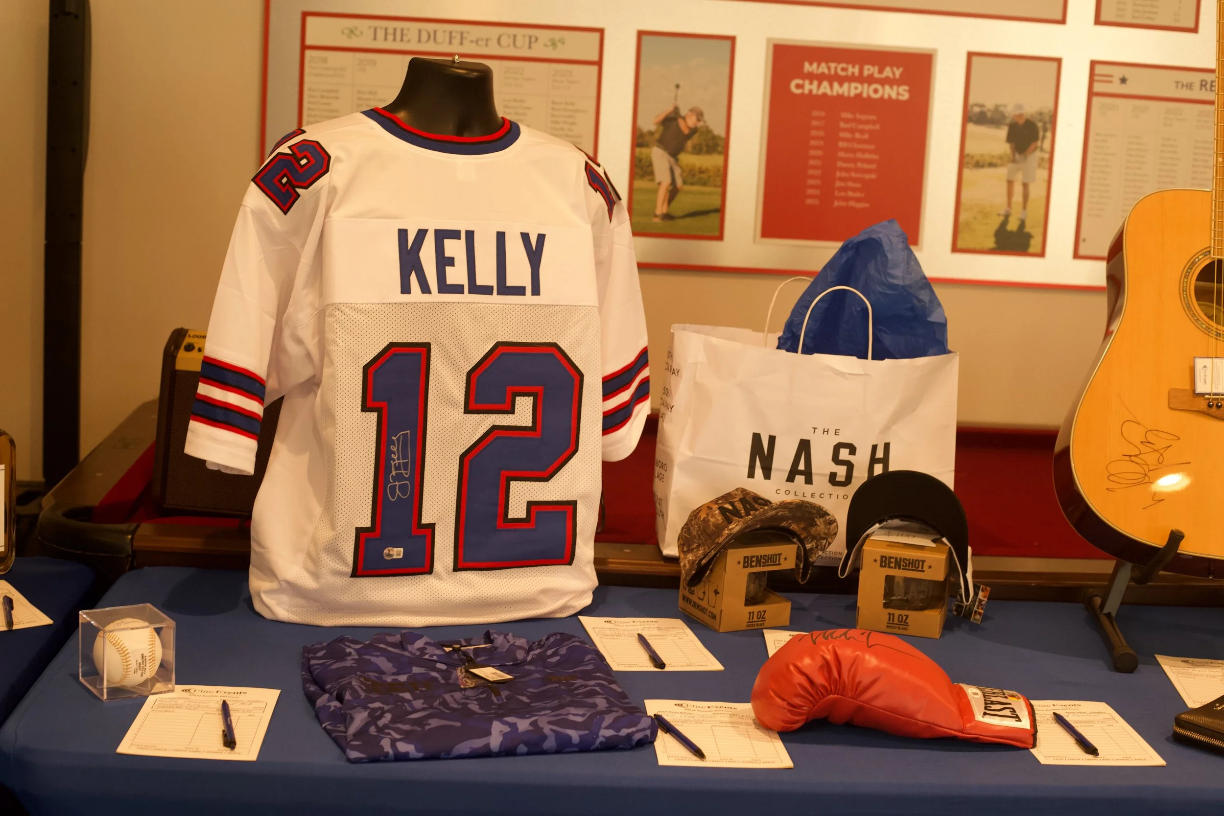 Display of sports memorabilia including a white hockey jersey with the name Kelly and number 12, a baseball bat, a baseball, a camouflage hat, a red boxing glove, and several pens and papers on a table, with memorabilia posters and photos in the back
