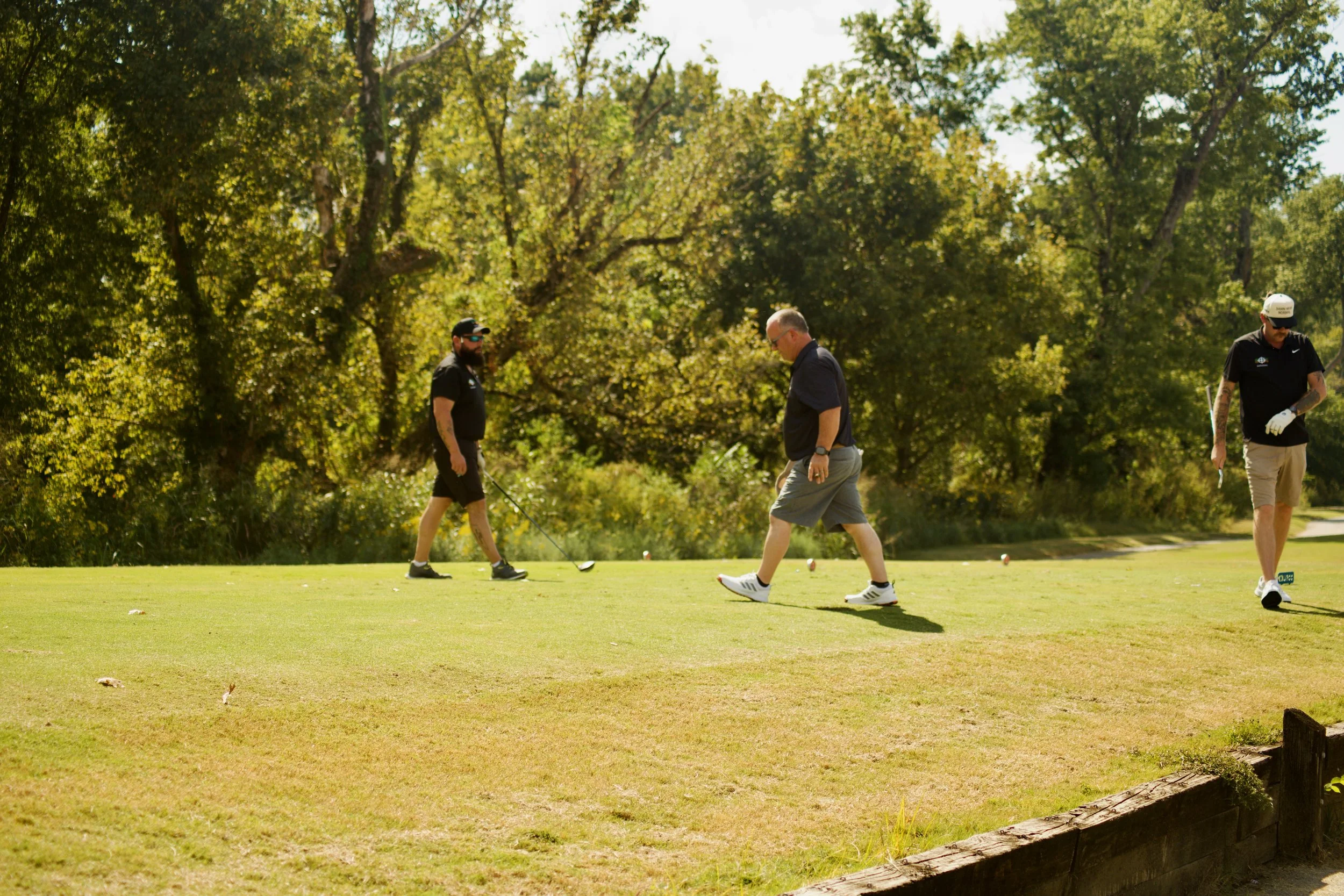 Three men playing or practicing golf on a sunny day in a park-like setting with trees in the background.