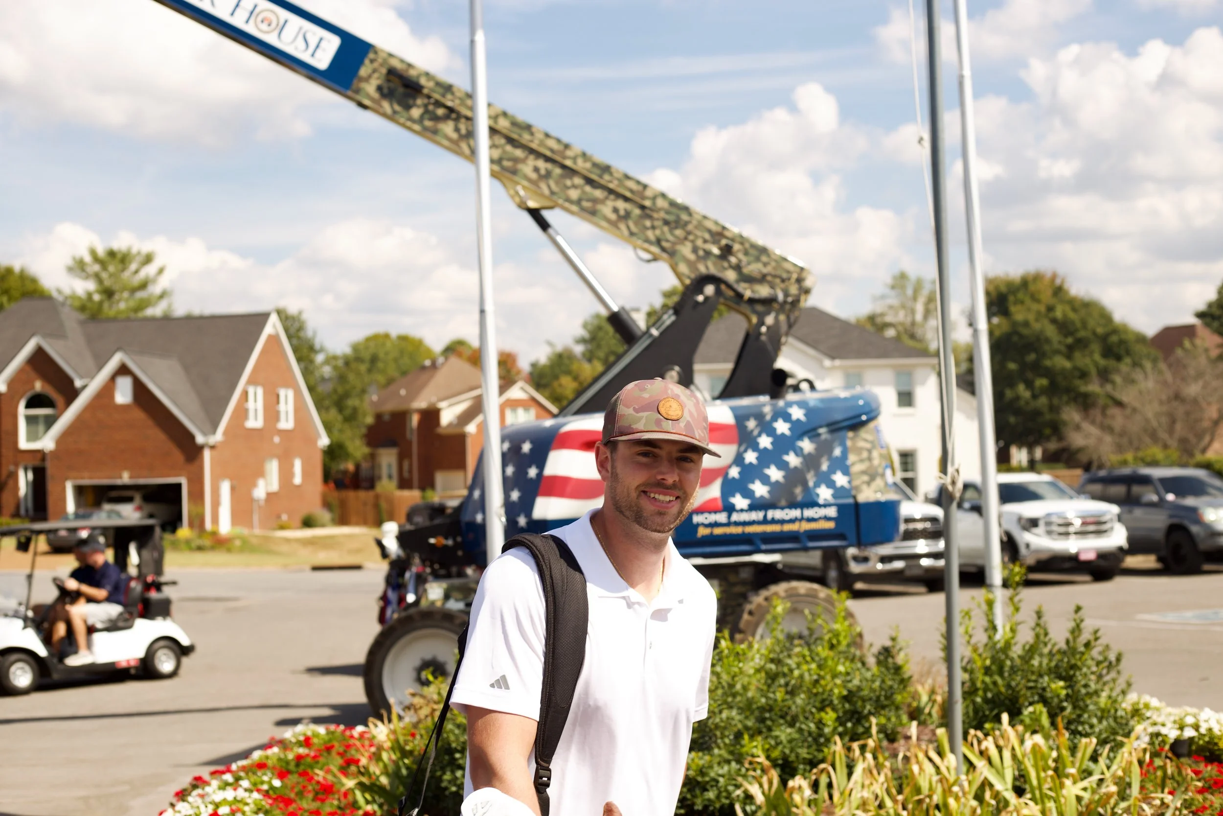 A young man wearing a white shirt and a camouflage cap stands outside at an event with a flight simulator in the background. The simulator is decorated with an American flag theme and a sign that reads "Home Away From Home." The setting is a resident
