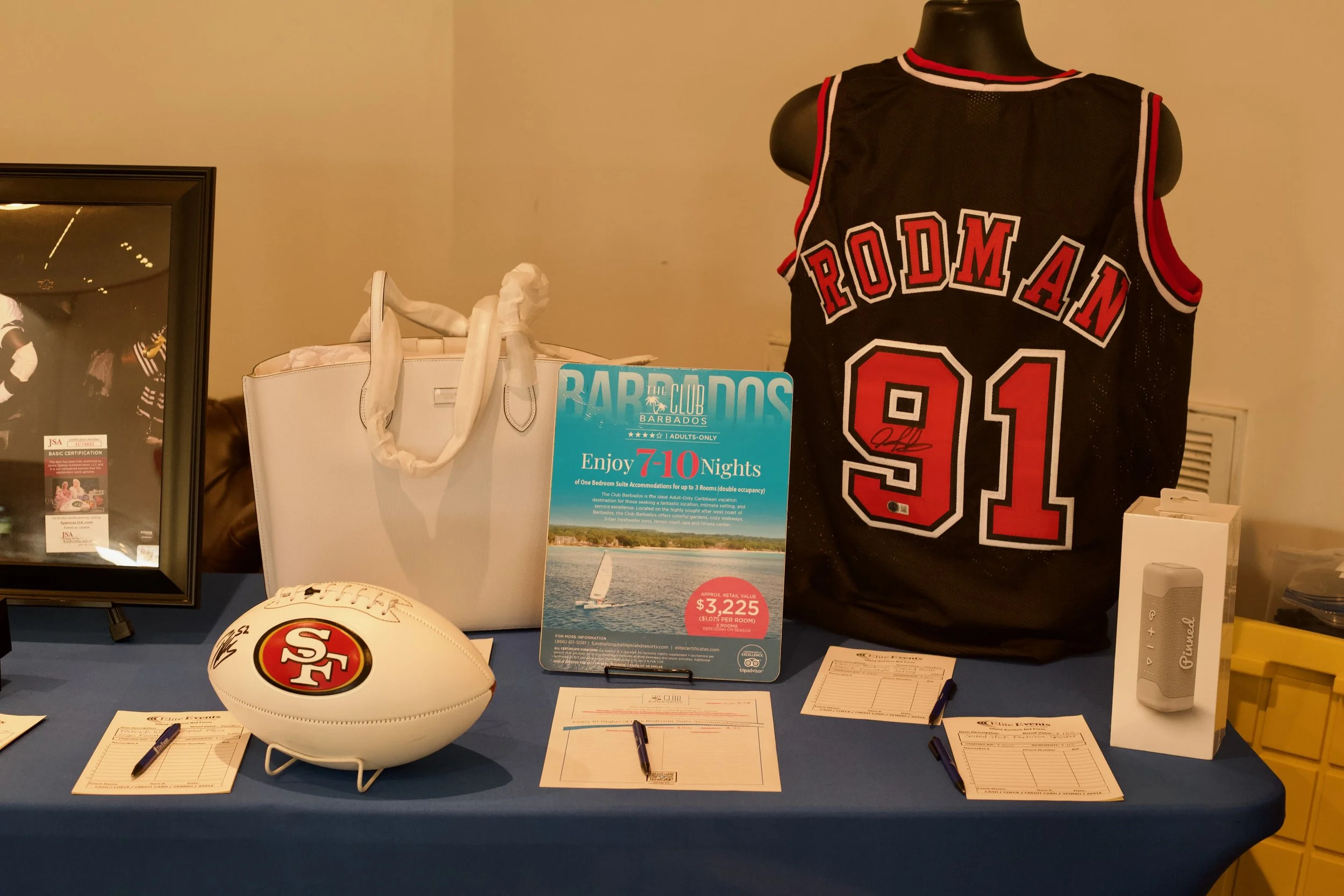 Display table with San Francisco 49ers football memorabilia, including an official football, a signed basketball jersey, and promotional flyers for vacations to Barbados.