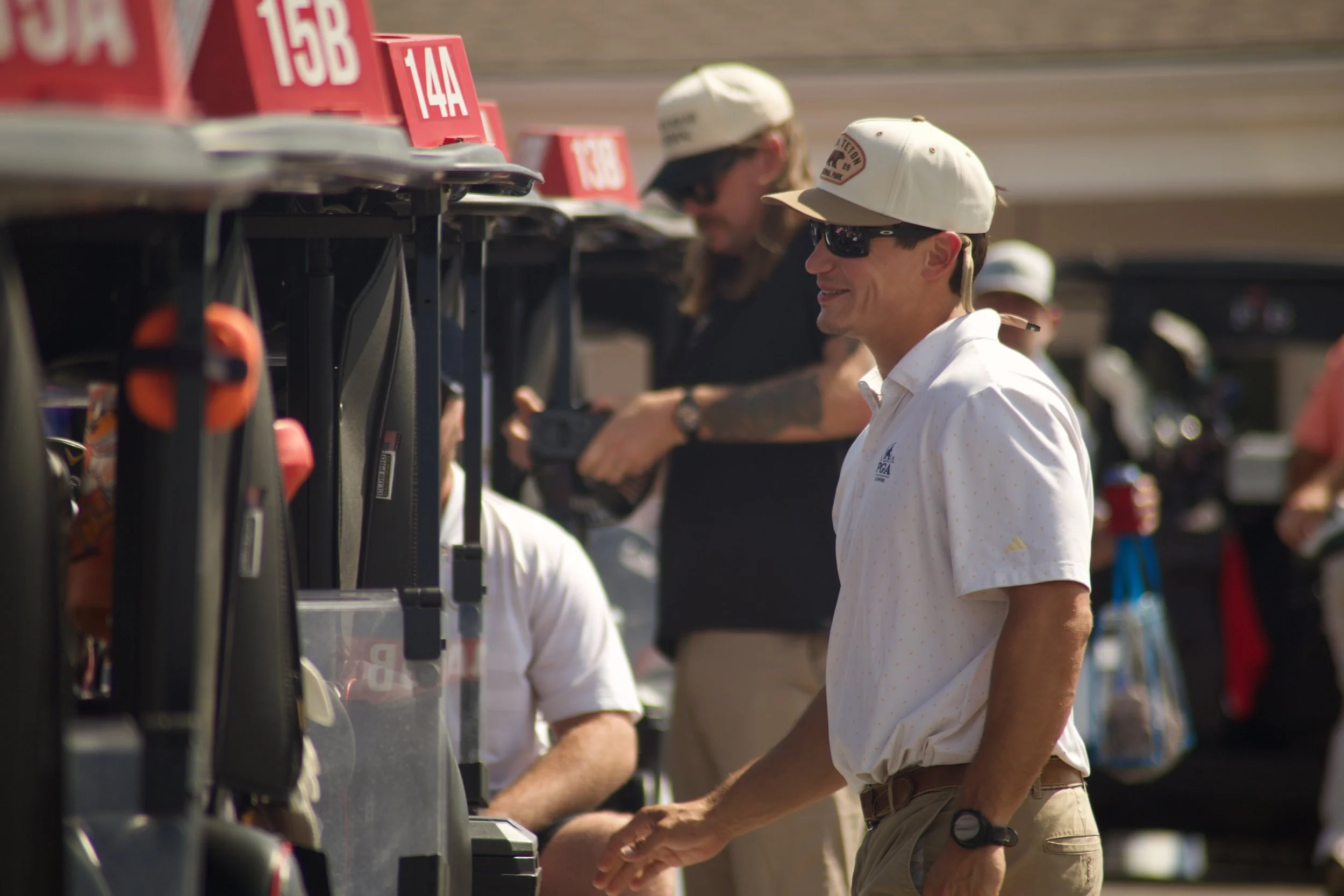 Two men at a golf course in bright sunlight. One is wearing a white cap, sunglasses, white polo shirt, and khaki shorts, and is smiling. The other man is wearing a white cap, sunglasses, and a black shirt. They are standing in front of a row of golf 