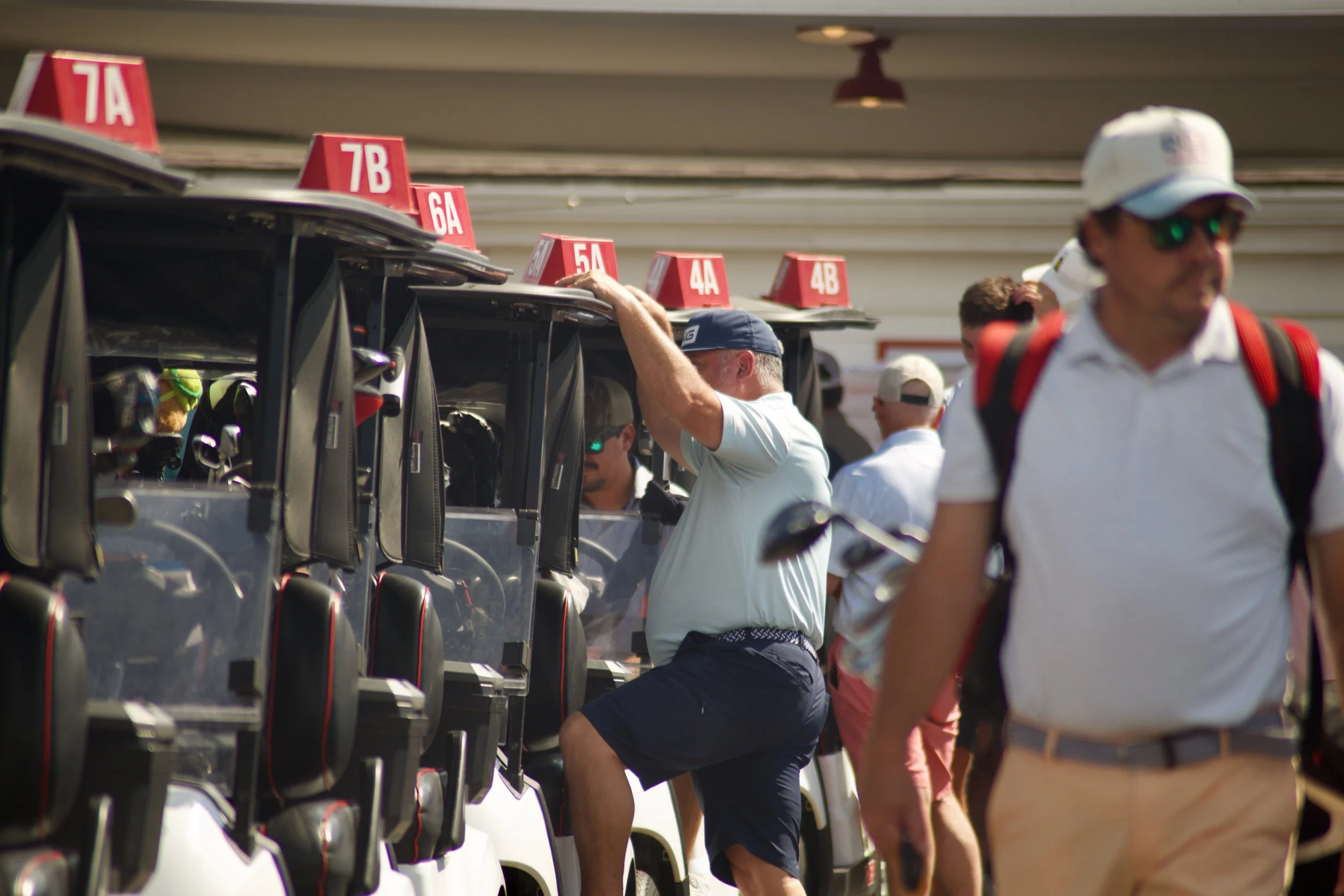 Golf carts lined up at a golf course with people walking beside them, some wearing caps and sunglasses, during daytime.