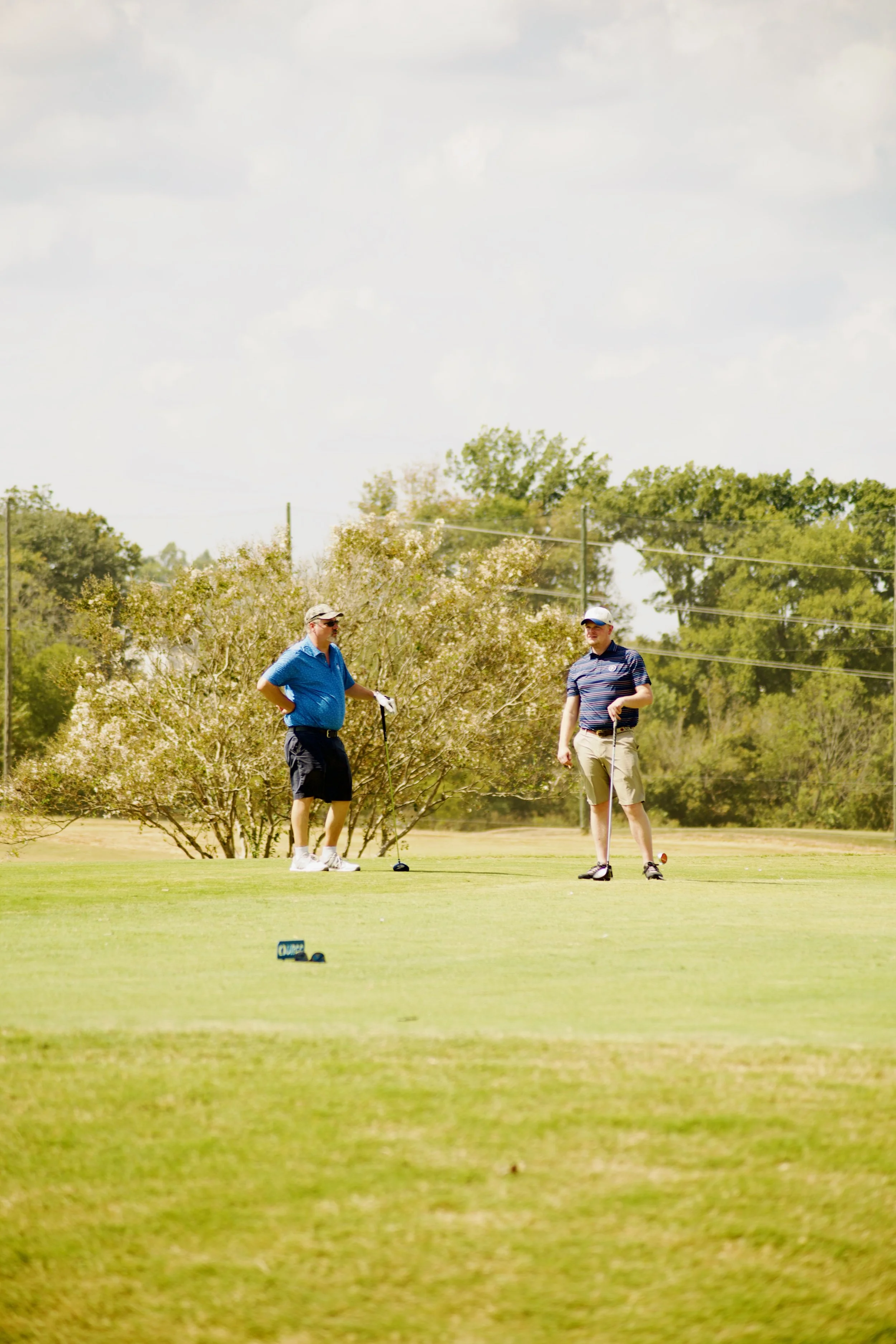 Two men playing golf on a sunny day on a golf course with trees and power lines in the background.