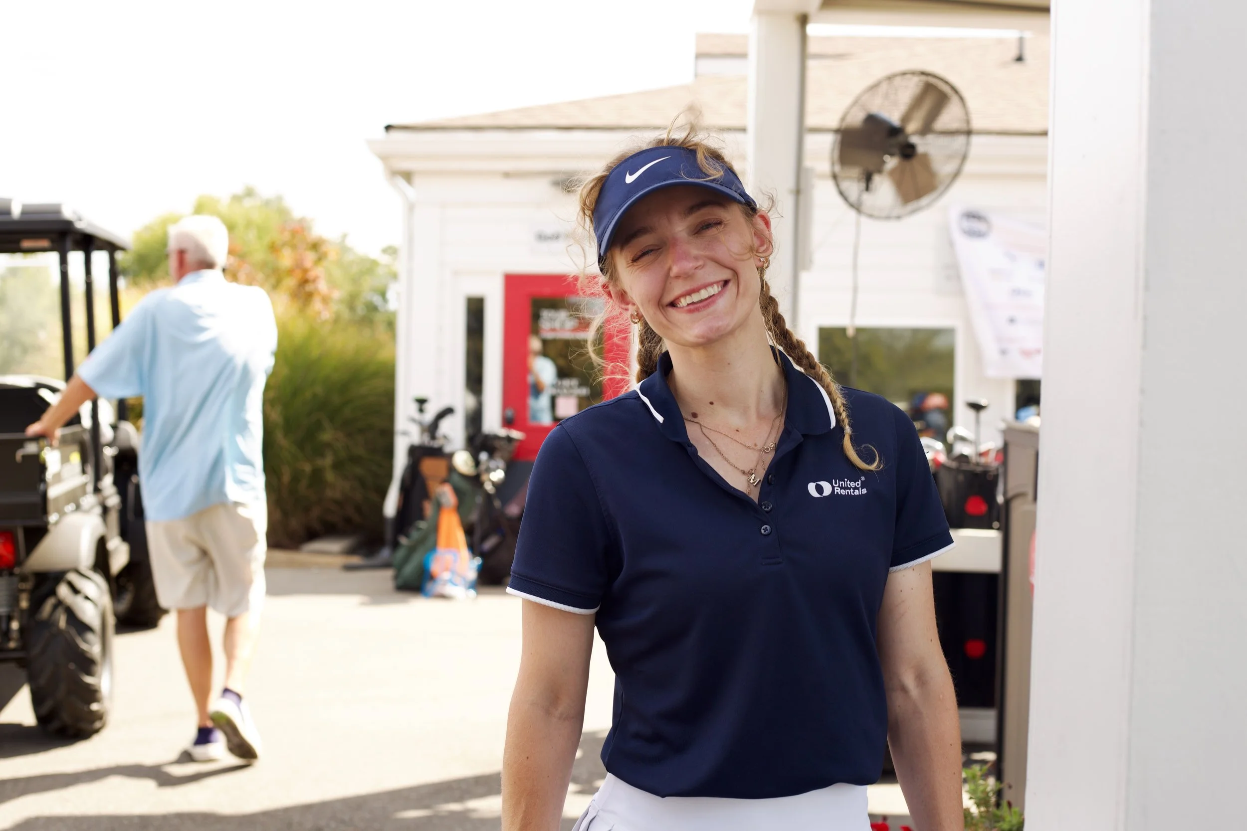 Smiling young woman in navy blue polo shirt and Nike visor stands outdoors with golf equipment and a golf cart in the background.