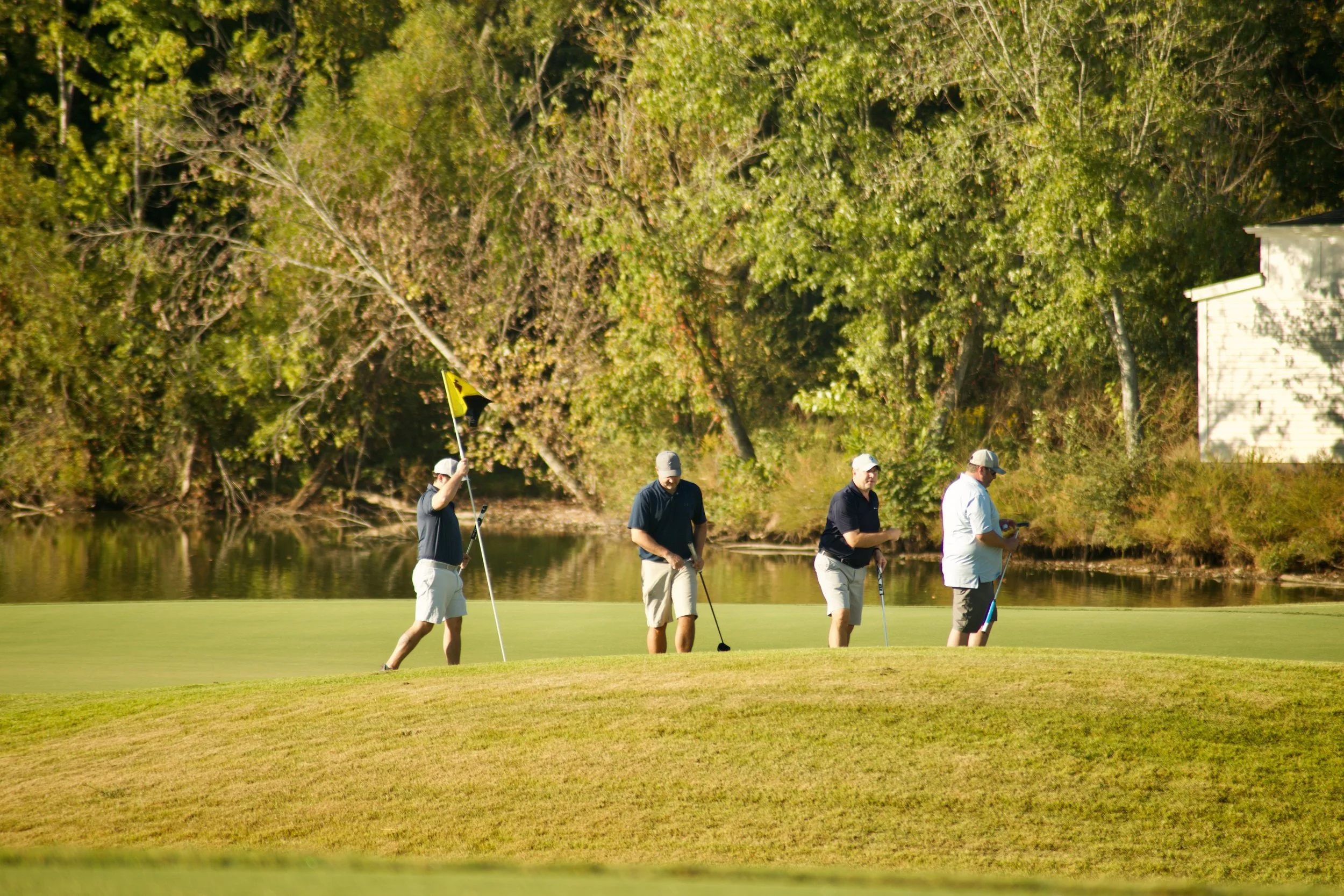 Four men dressed in golf attire walking on a golf course near water, with trees and a white building in the background.