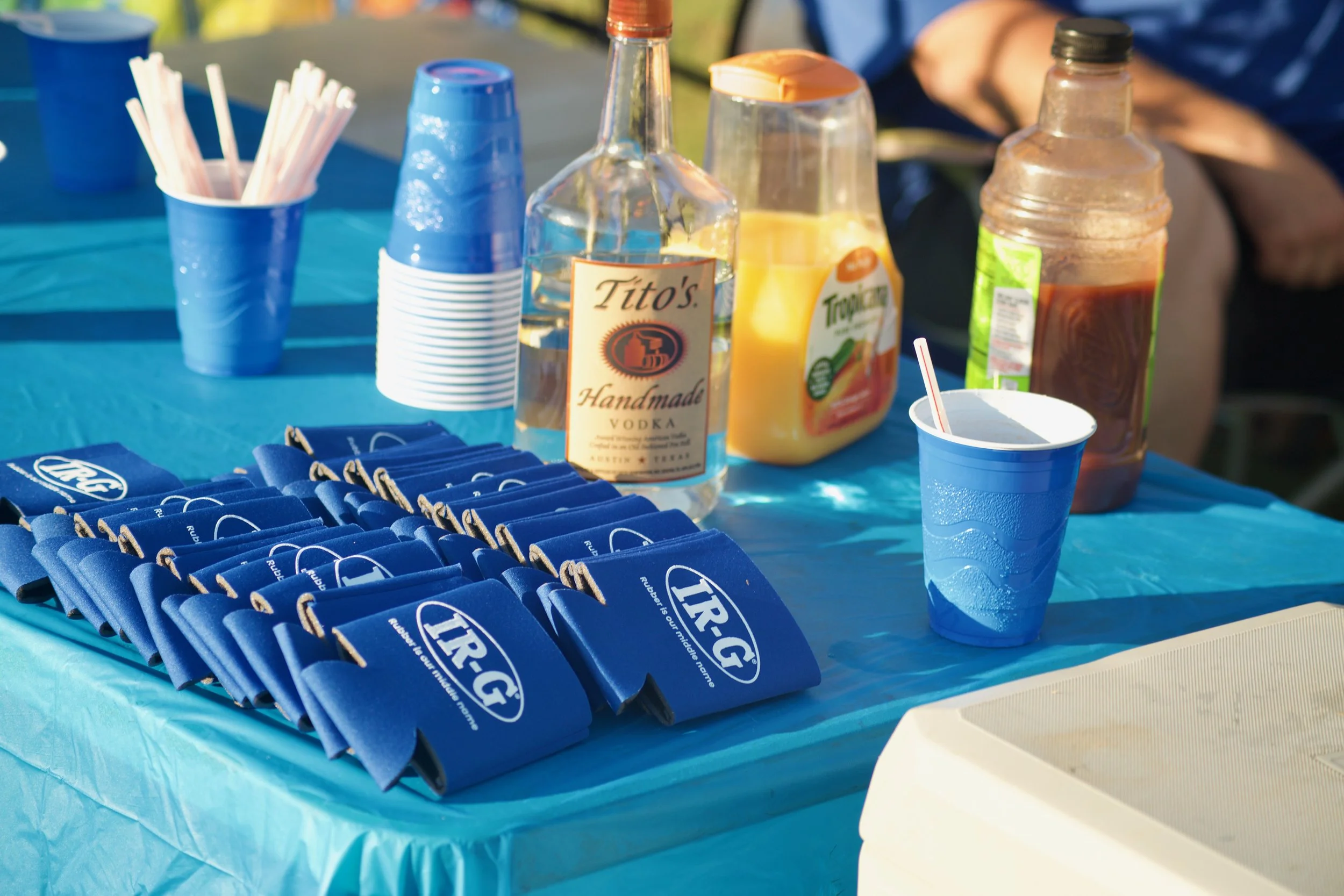 A table with blue cups, disposable straws, and various condiment bottles including Tito's handmade vodka, Tropicana orange juice, and a hot sauce bottle, likely set up for an outdoor event or gathering.