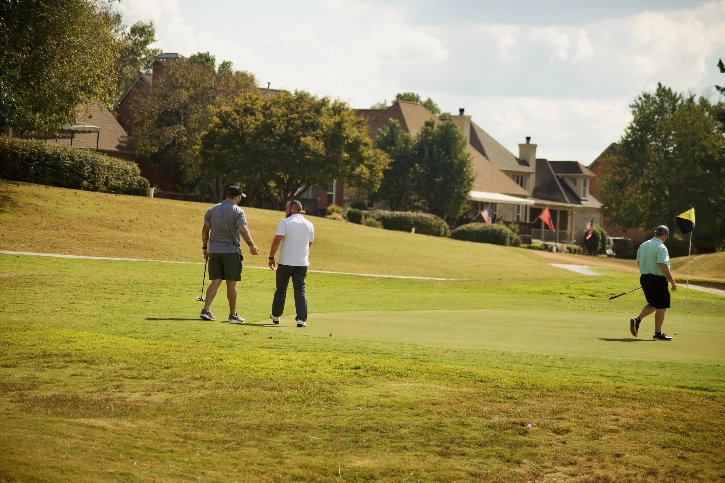 Three men playing golf on a sunny day at a golf course, with houses and trees in the background.