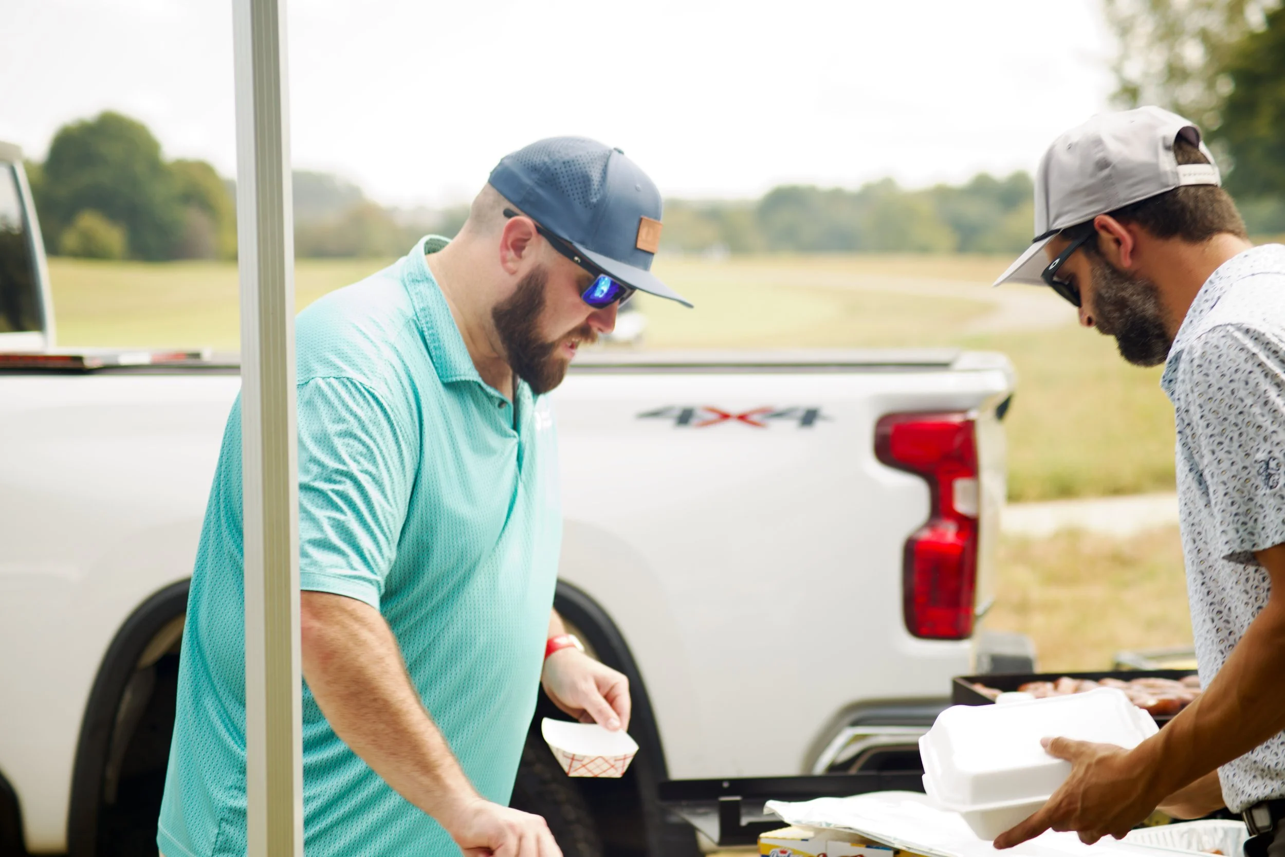 Two men are at an outdoor barbecue. One man, with a beard and wearing a blue cap and light blue shirt, is holding a small paper food tray. The other man, with a beard, sunglasses, and a white cap worn backwards, is serving food from a container. A wh