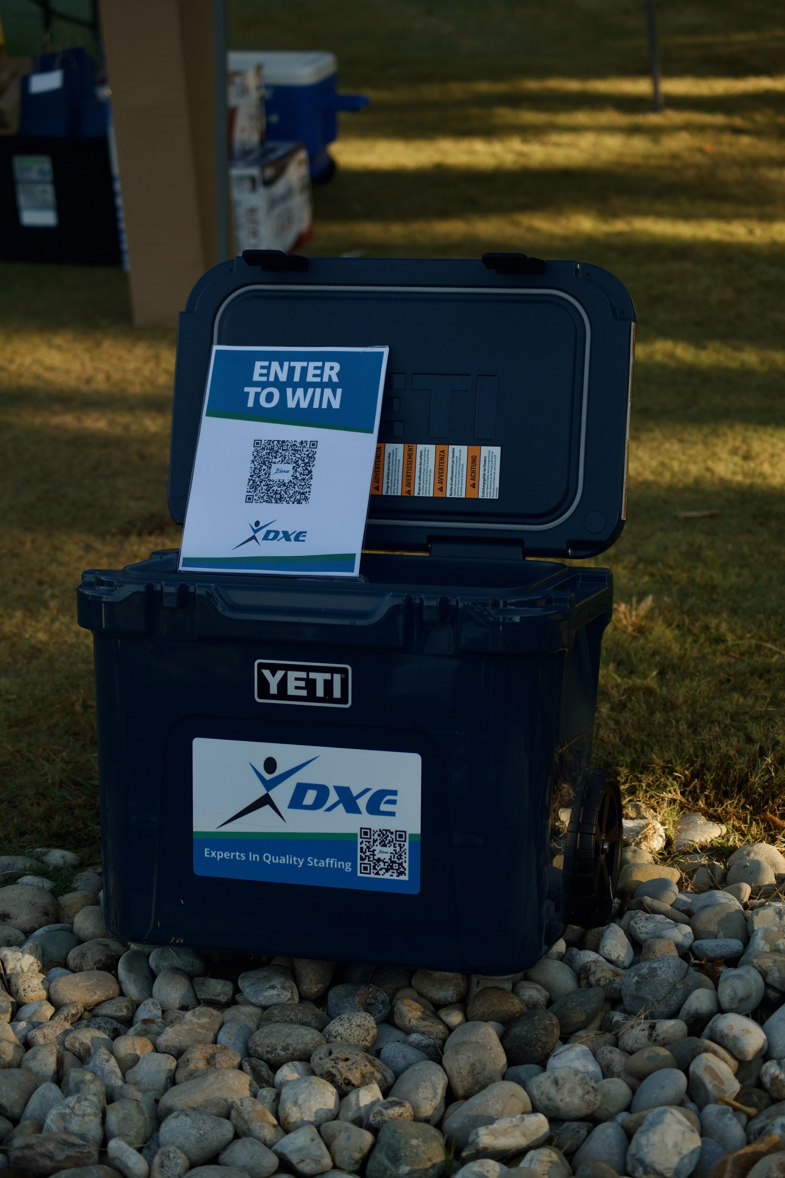 A black YETI cooler with a DXE promotional sign attached, displayed outdoors on a rocky surface with grass and a shadowed background.