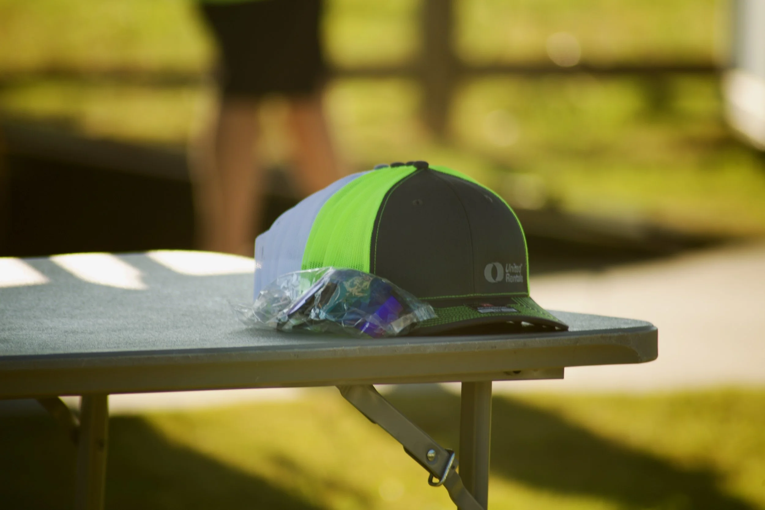 A gray and green baseball cap with the Union Rent logo resting on a table, with a small plastic bag containing sunglasses or eyeglasses beside it, under natural outdoor lighting.