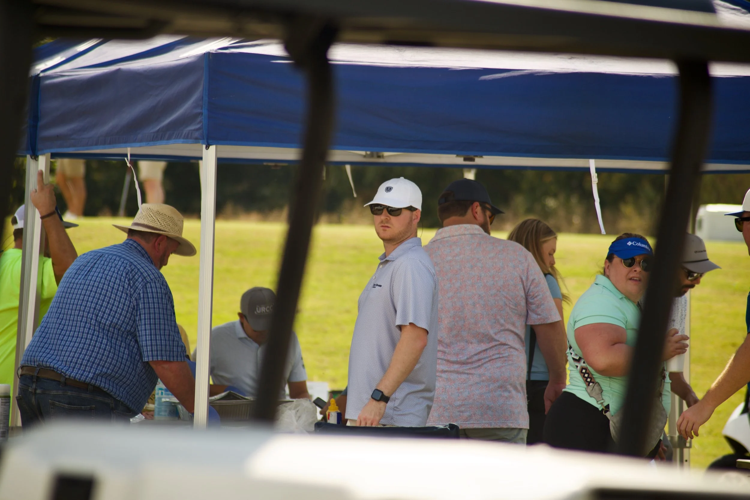 People gathered under a blue canopy outdoors, some are standing near a table with items, and there are trees in the background.