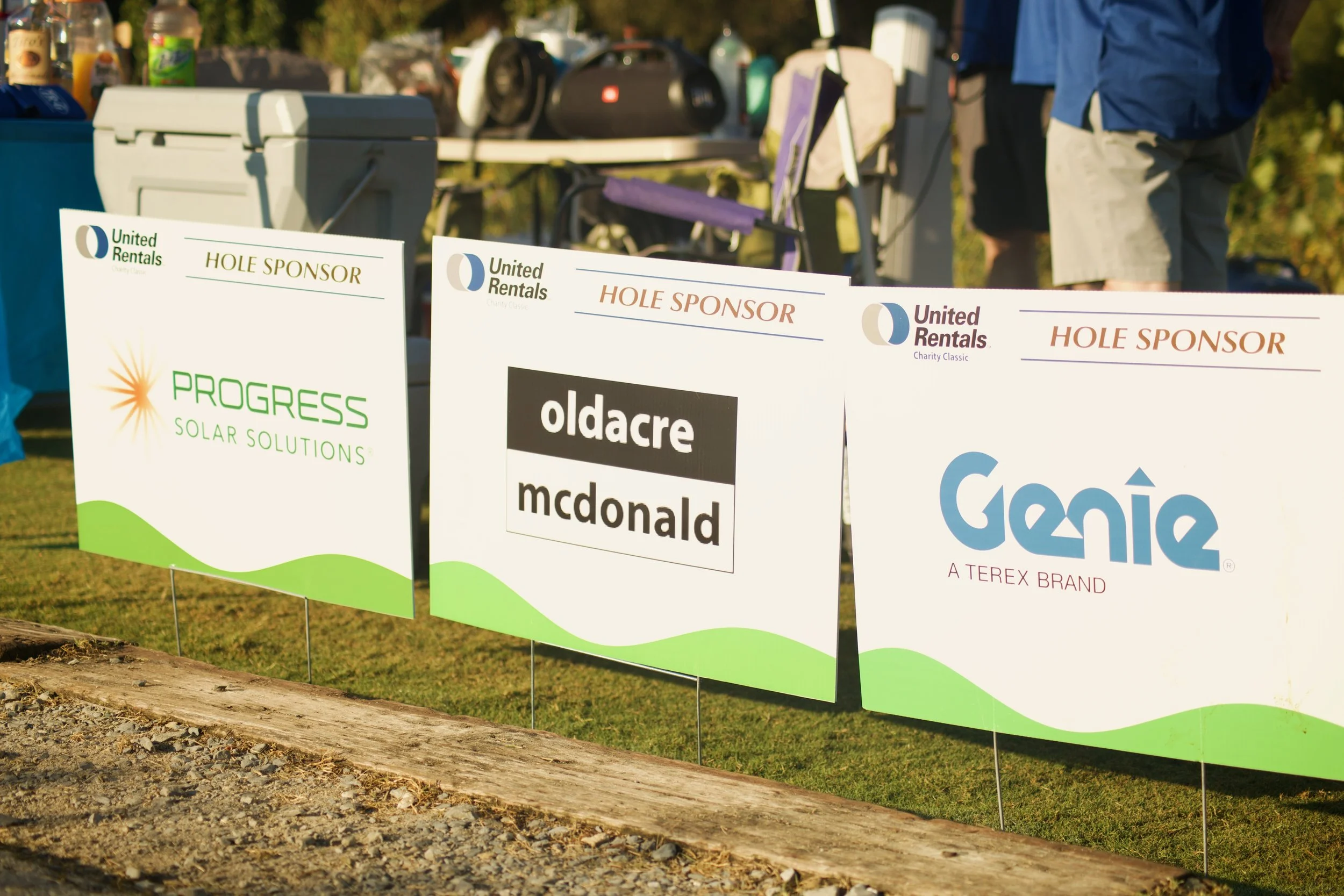 Several signs promoting sponsors for a golf event, including 'Progress Solar Solutions,' 'Oldacre McDonald,' and 'Google,' displayed on a fenced boundary with a grassy area and trees in the background.