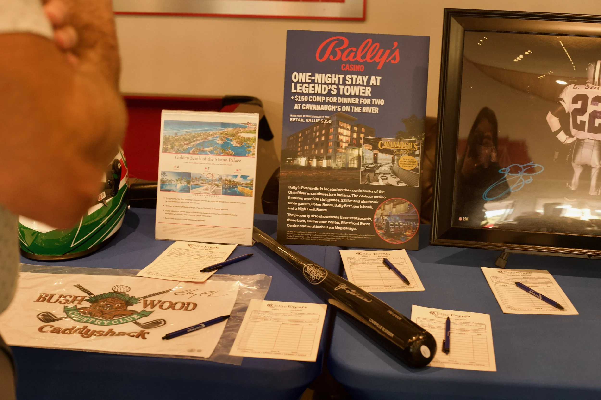 Table display at a promotional event featuring Bally's Casino with promotional flyers, a framed photo of a football player, a black baseball bat, pens, and various informational brochures and sign-up sheets.