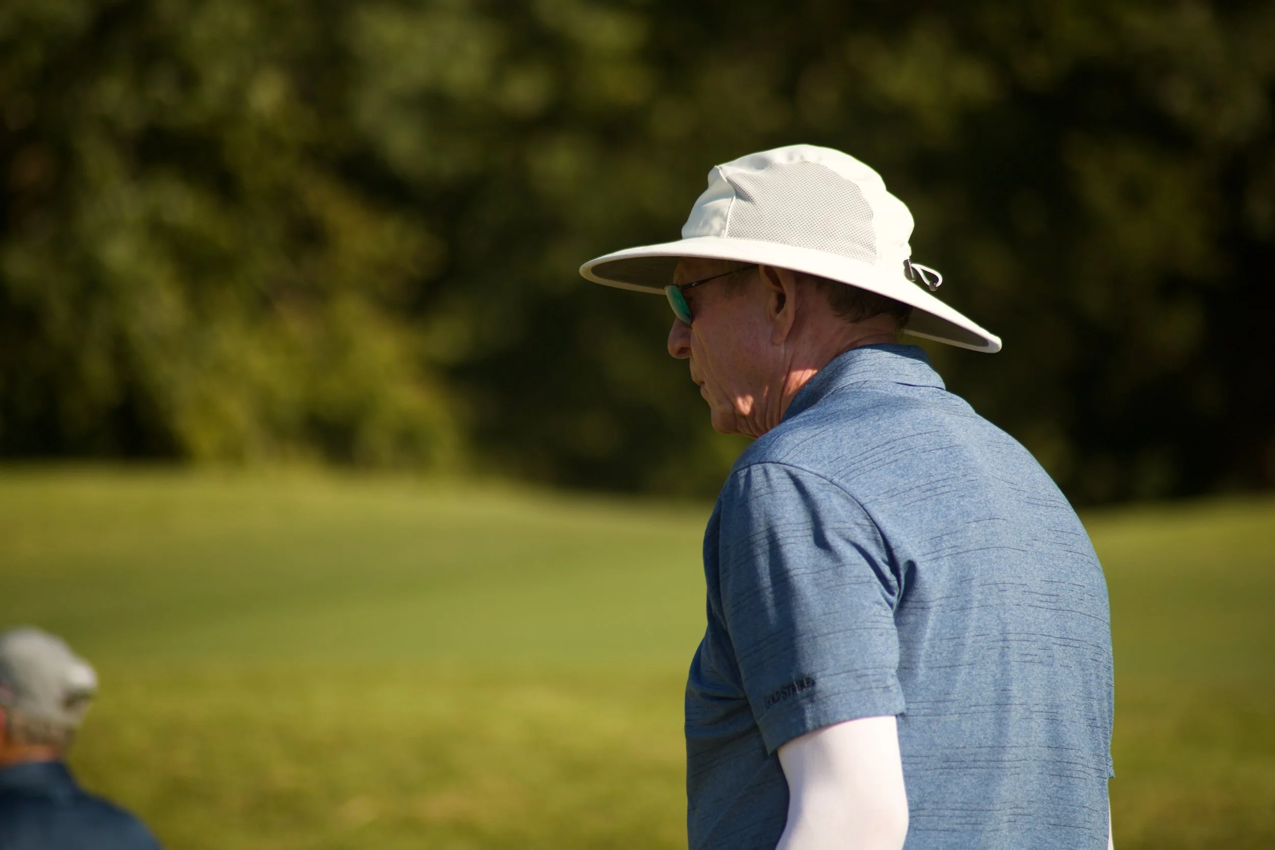 A person wearing a white wide-brimmed hat, sunglasses, and a blue polo shirt, sitting outdoors on a sunny day with a blurred green natural background.