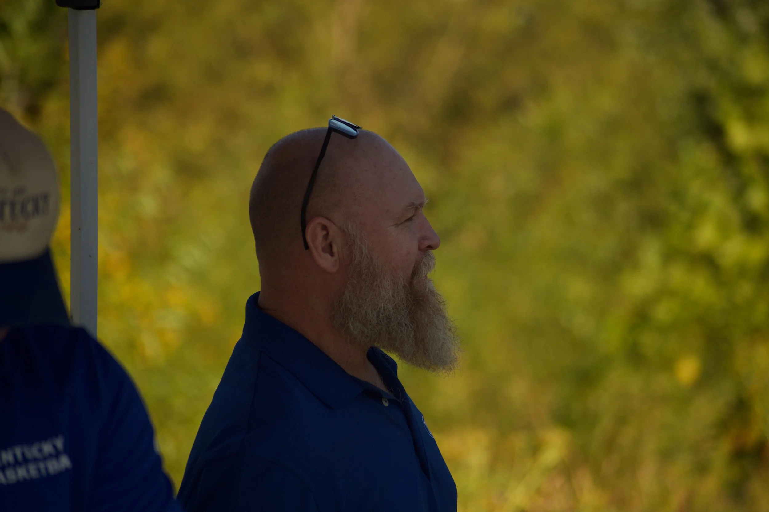 Profile of a bald man with a beard wearing sunglasses on his head and a navy blue polo shirt, standing outdoors against a background of blurred green and yellow foliage.