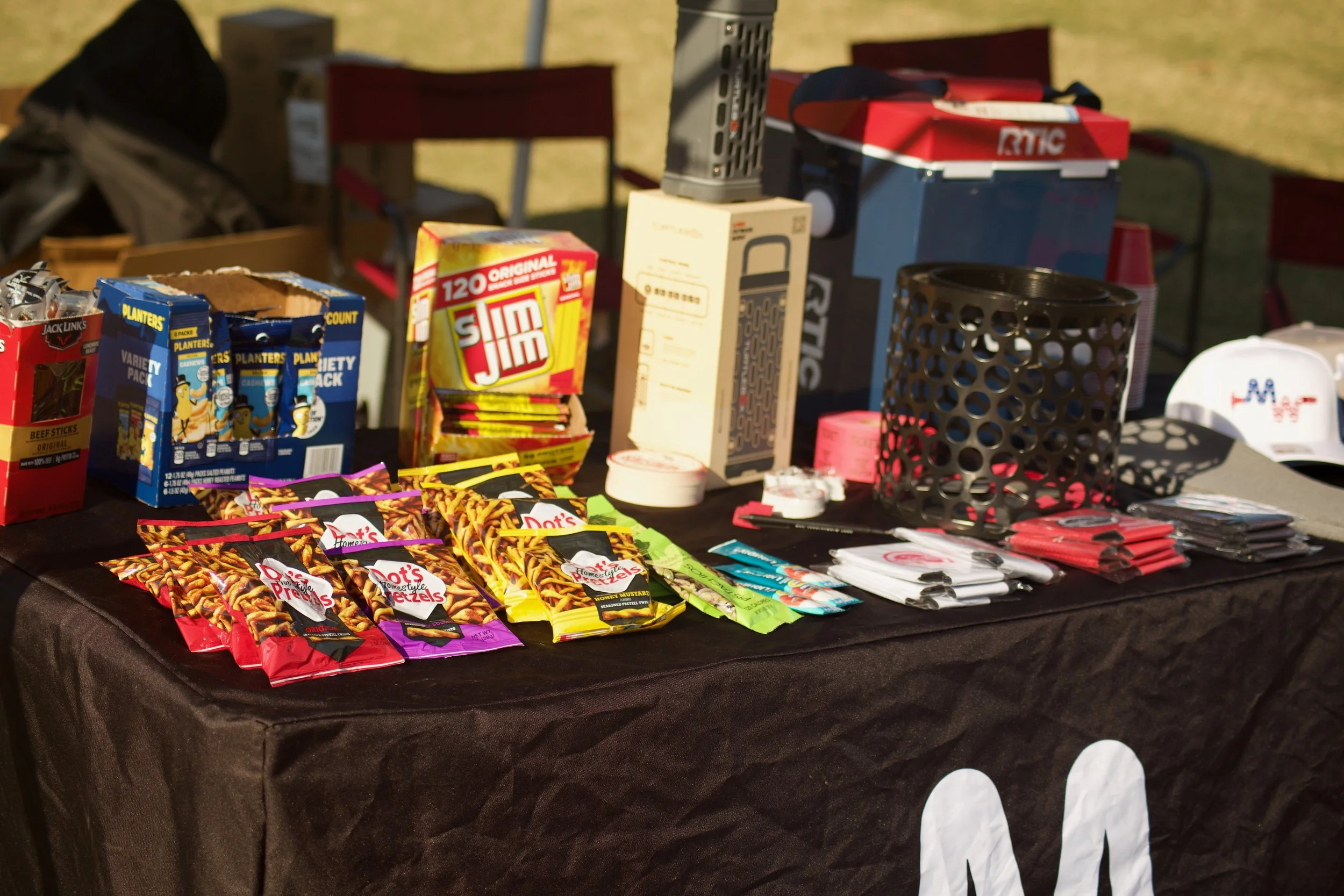 A table covered with a black tablecloth displaying various snack foods, small giveaway items, and promotional materials at an outdoor event.