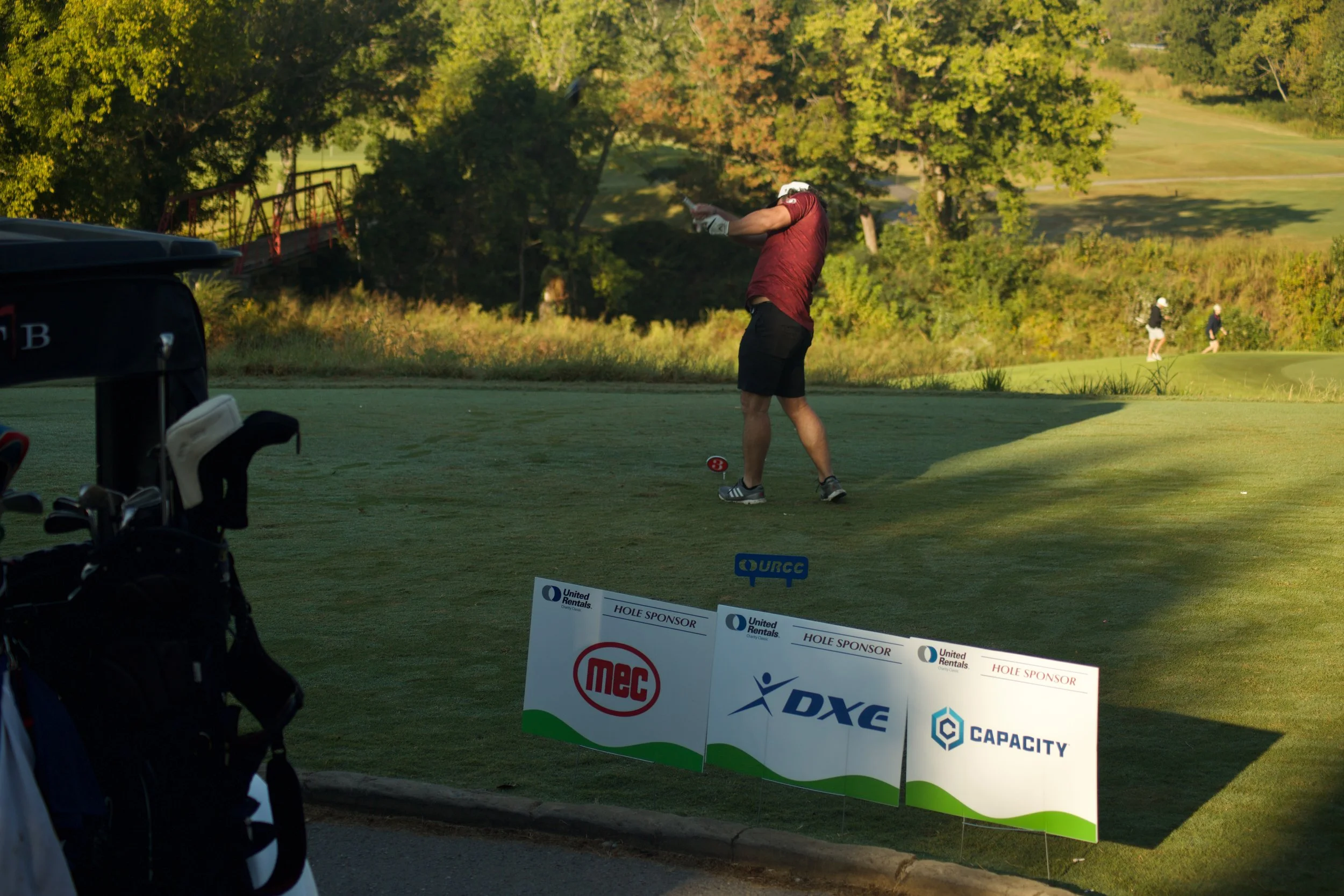 A male golfer in a red shirt and black shorts is swinging a golf club on a golf course surrounded by green trees and grass. In the foreground, there is a golf bag and a sponsor sign with various logos.