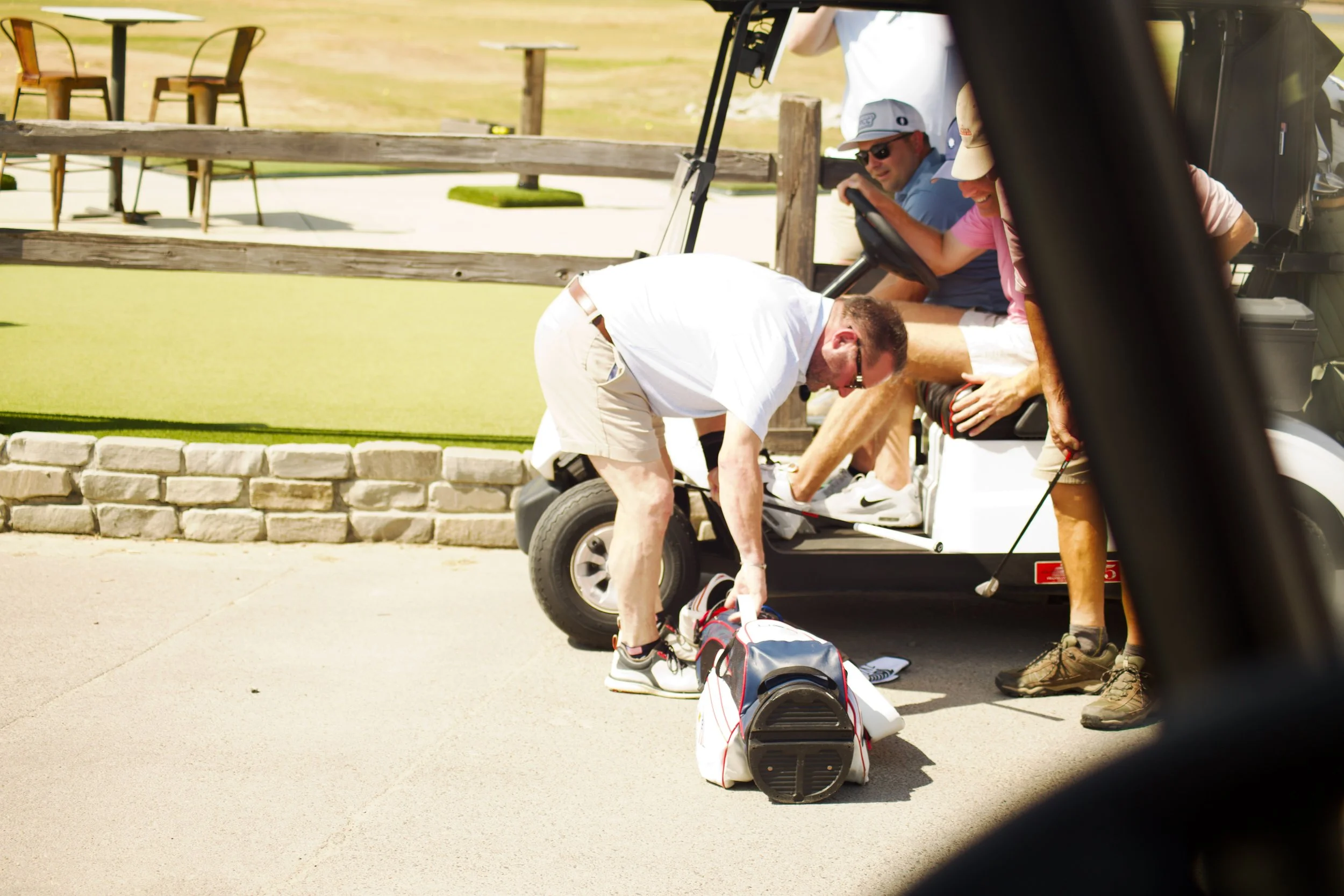 A group of people at a golf course, sitting in a golf cart. One person is standing outside the cart, tying or adjusting their golf shoes, while others are seated inside. The scene takes place on a sunny day.