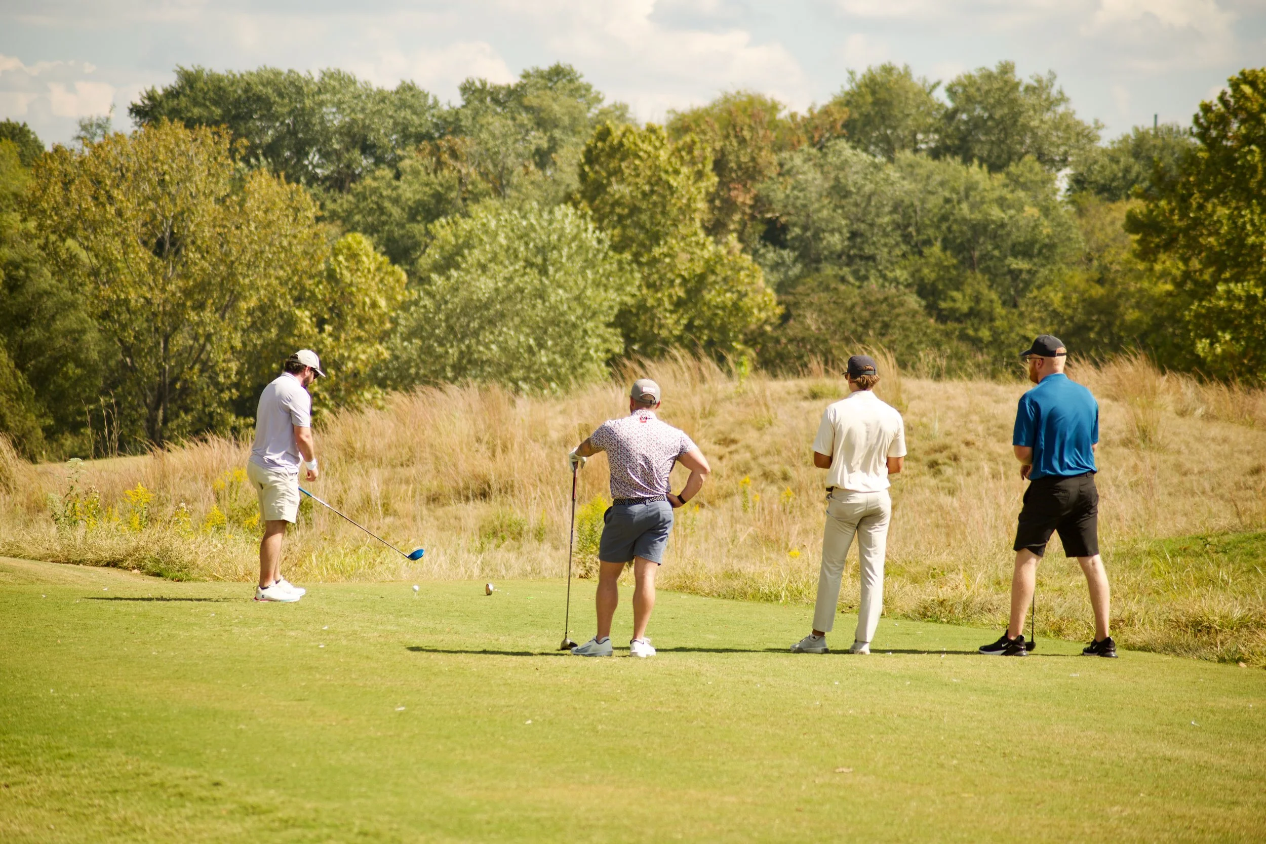 Group of five men playing golf on a lush green course with trees in the background under a partly cloudy sky.