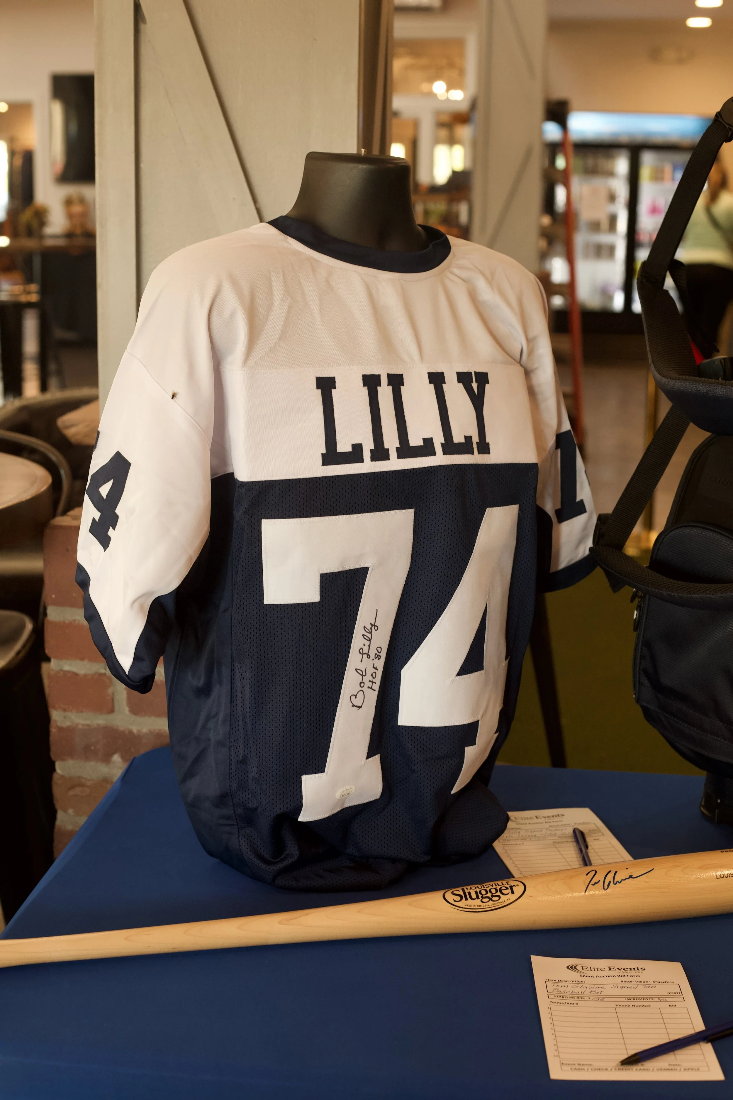 A mannequin wearing a football jersey with the name 'LILLY' and the number '24' displayed next to a Louisville Slugger baseball bat and an auction bid sheet on a blue table inside a restaurant or cafe.