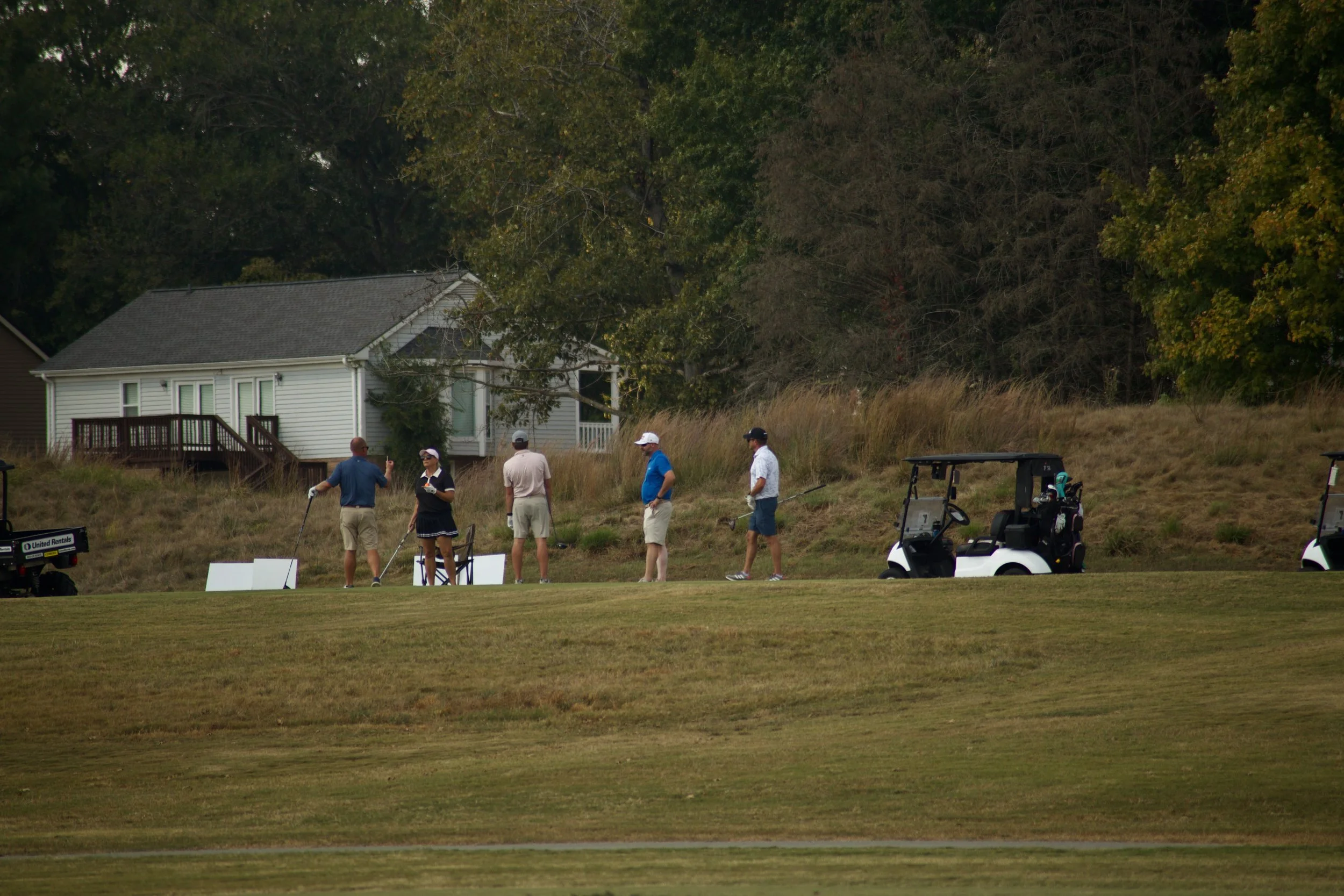 A group of people standing on a golf course, with golf carts nearby, and a house and trees in the background.