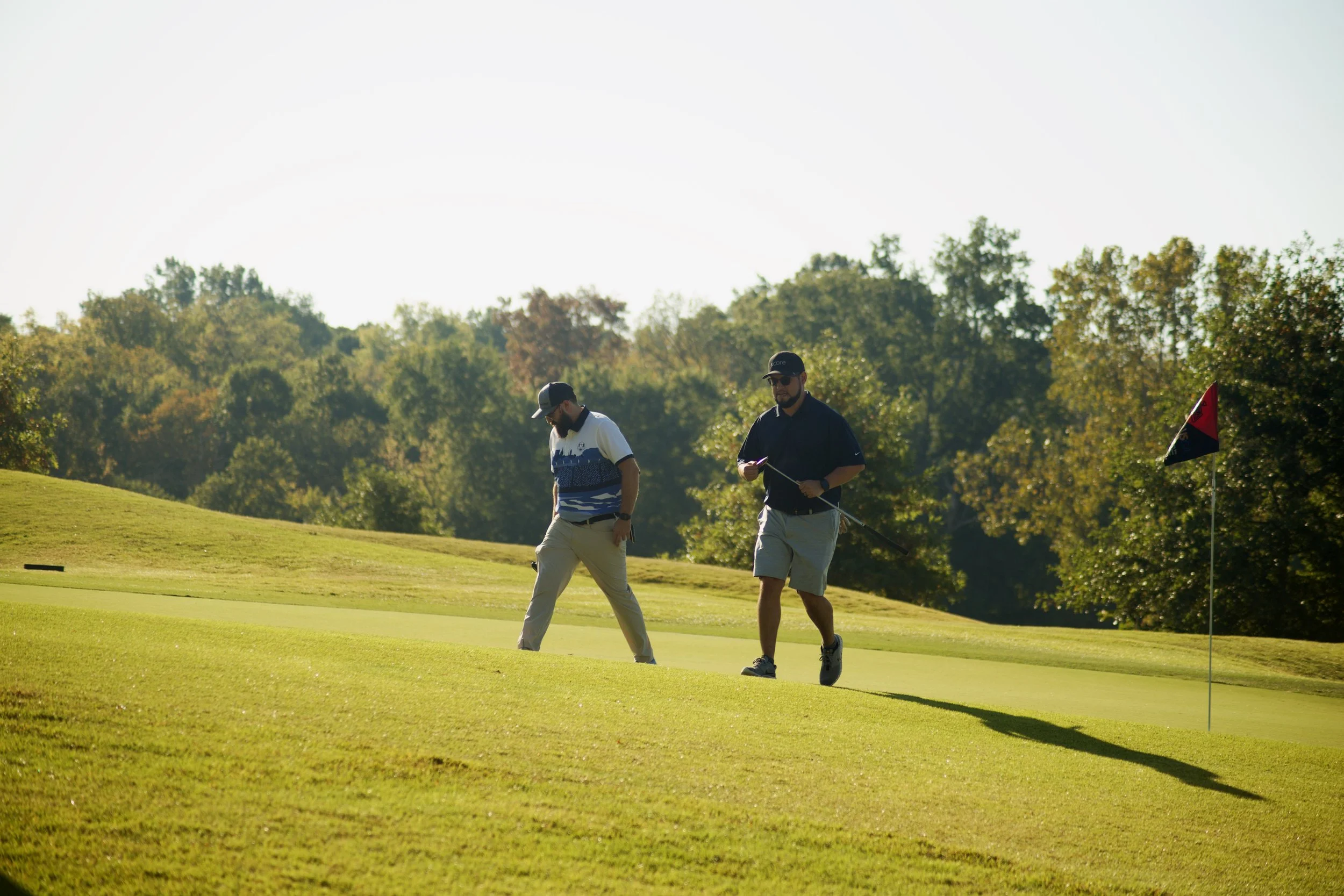 Two men playing golf on a sunny day near a flagstick on the green, with trees in the background.