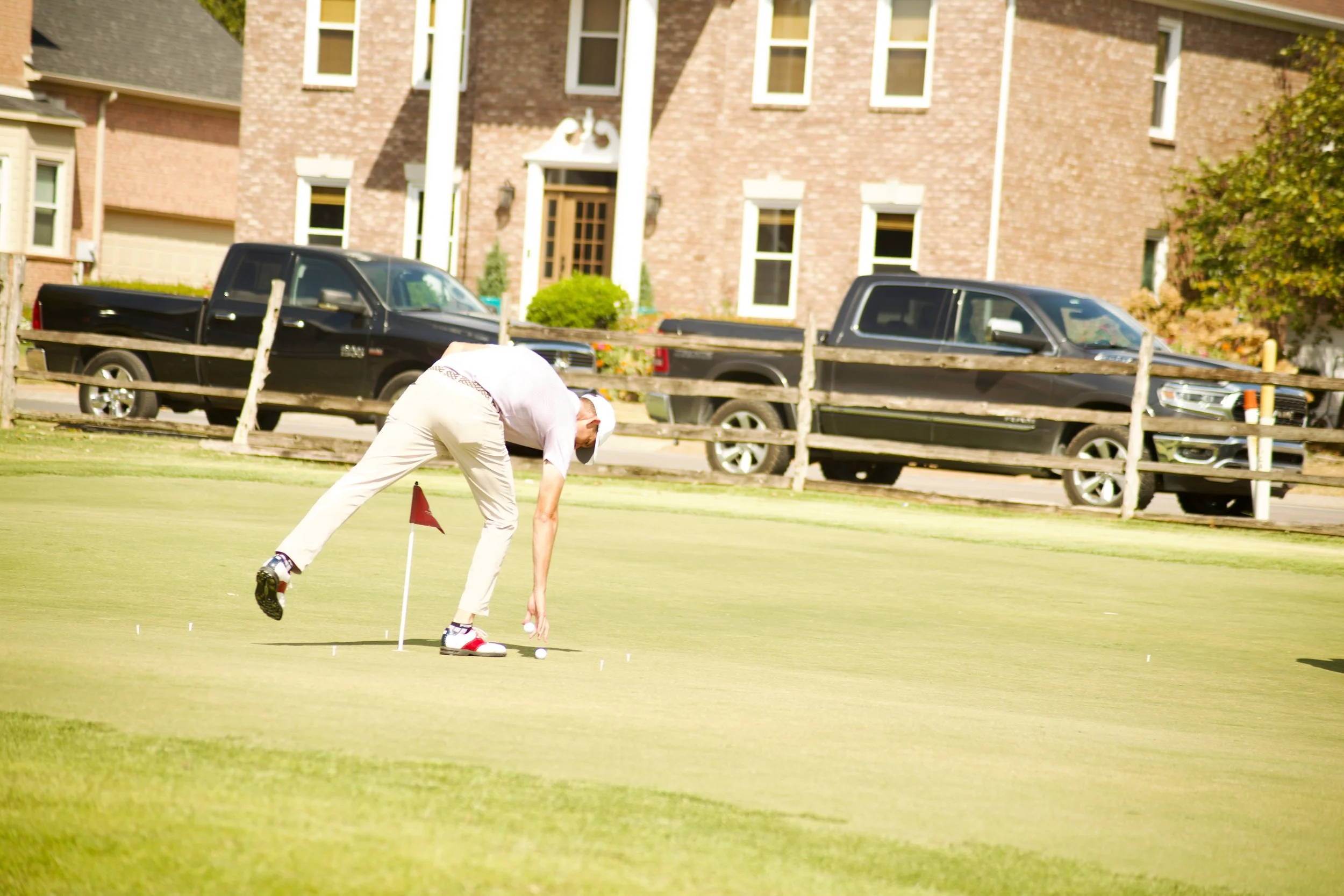 A man with a white cap on a golf course, bent over and examining the golf ball near the hole, with a wooden fence, two black trucks, and a brick house with multiple windows in the background.